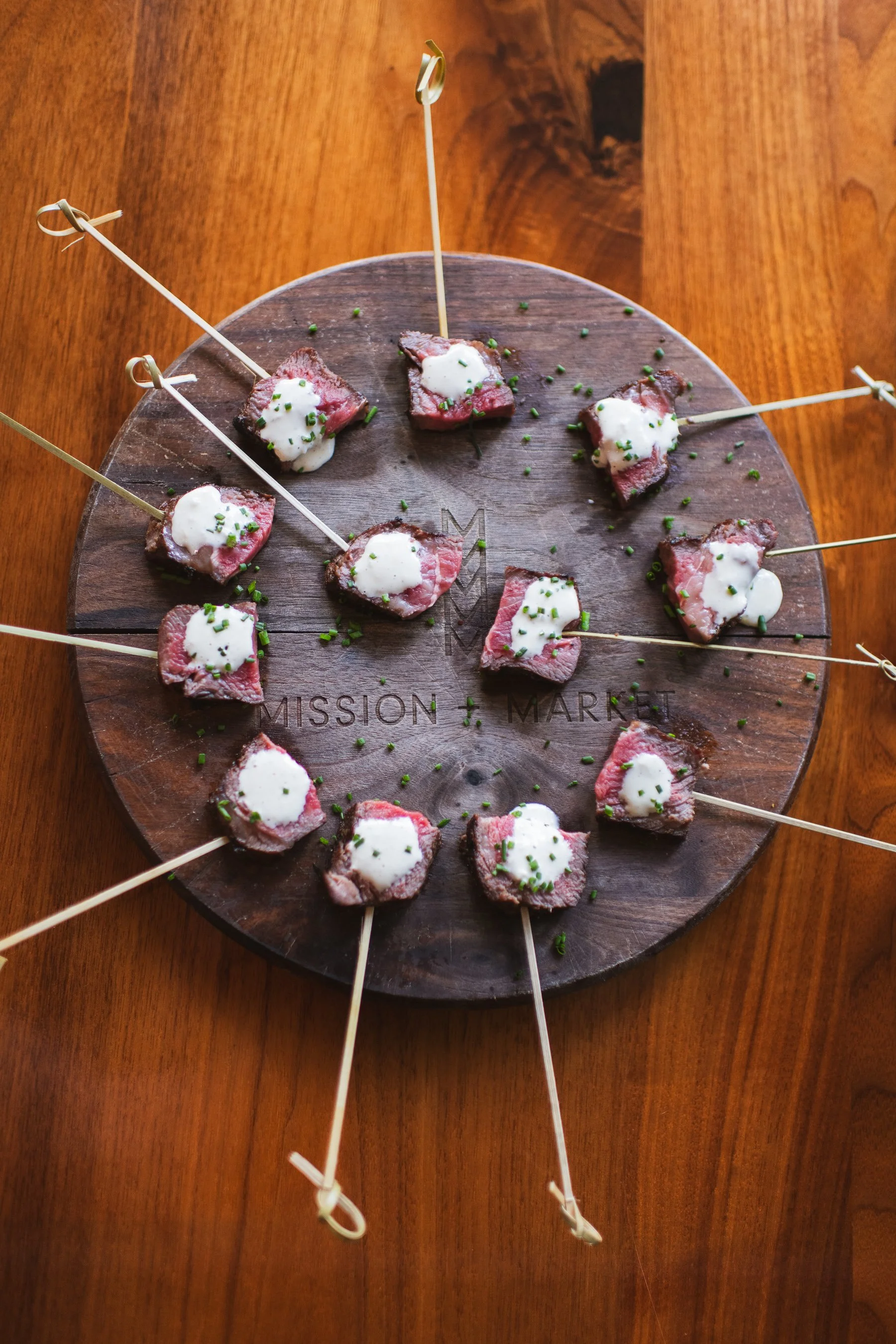 Slices of steak topped with a sauce and chopped chives, served on skewers, arranged on a round wooden board.