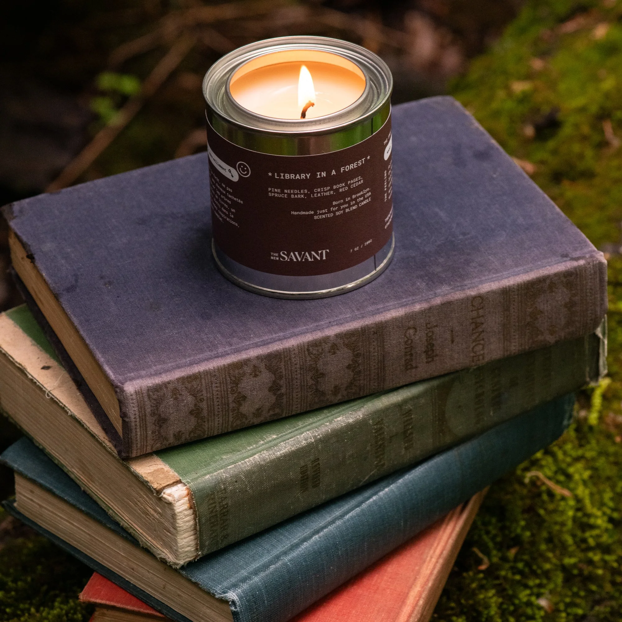 A candle in a metal can with a label, placed on a stack of old, weathered hardcover books outdoors. The candle is lit, casting a warm glow.