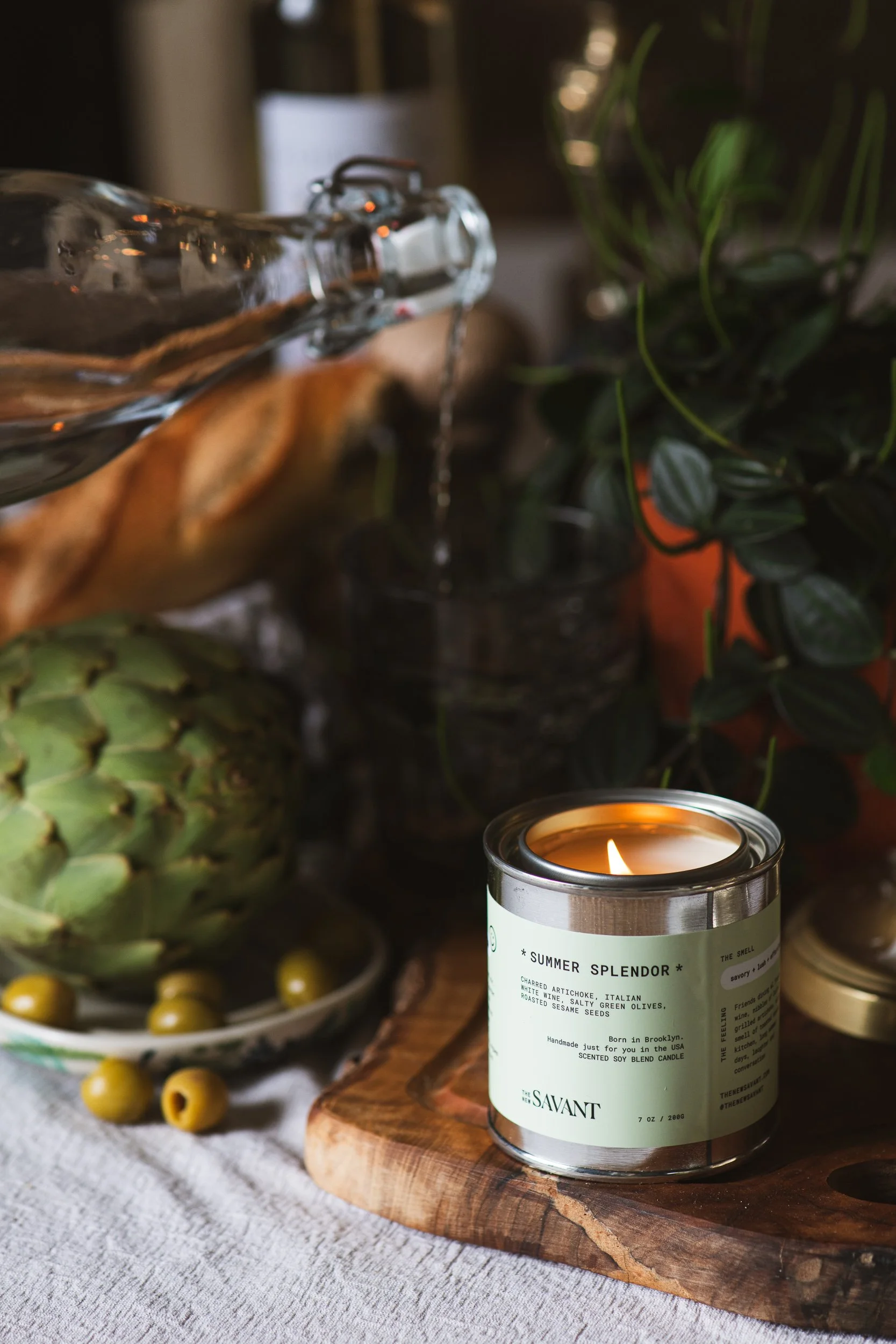 A lit candle labeled 'Summer Splendor' on a wooden surface, surrounded by decorative vegetables including artichoke and green olives, with plants in the background.