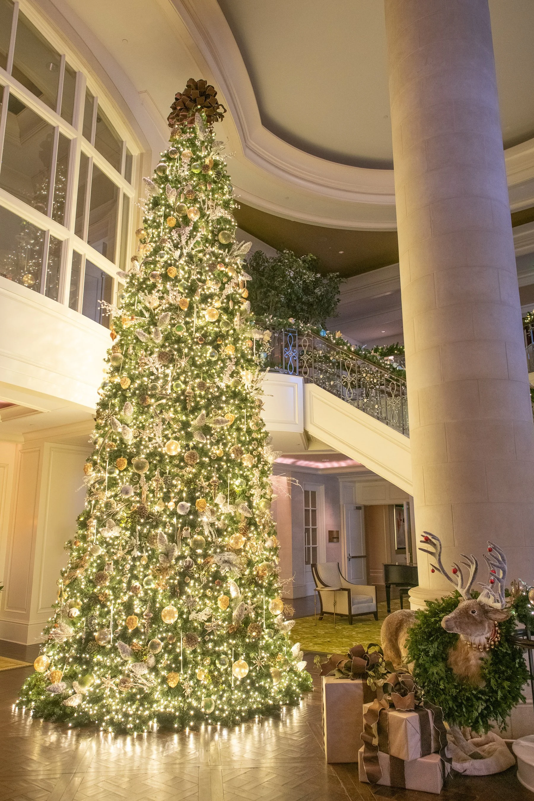 St. Regis Atlanta Christmas Decorations. A decorated Christmas tree with lights and ornaments in a large, elegant hotel lobby. To the right, there are wrapped gifts and reindeer decorations.