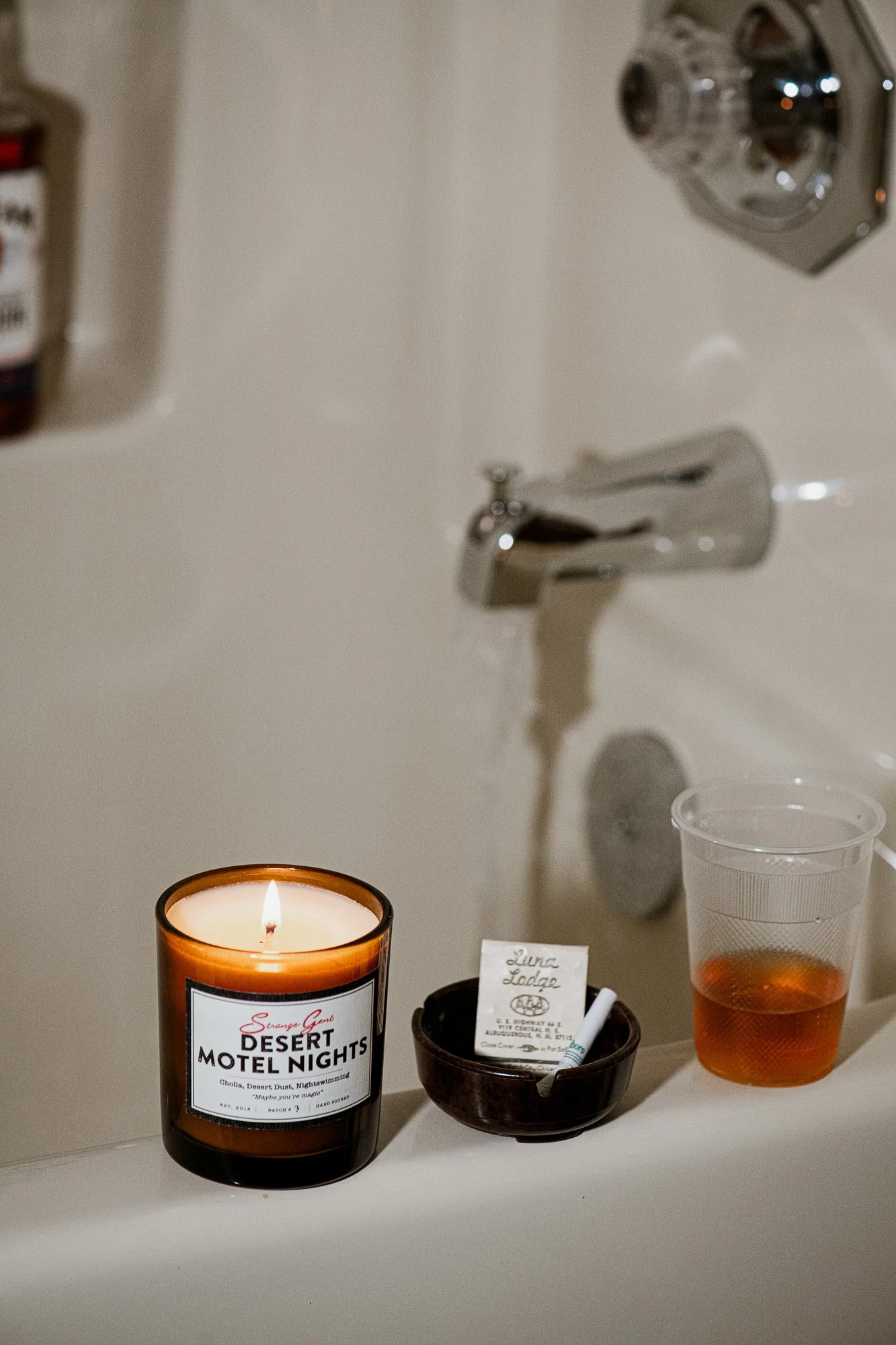 A lit candle with a label reading 'Desert Motel Nights' on a bathroom sink, alongside a small container with a business card, a pen, and a plastic cup filled with amber-colored liquid.