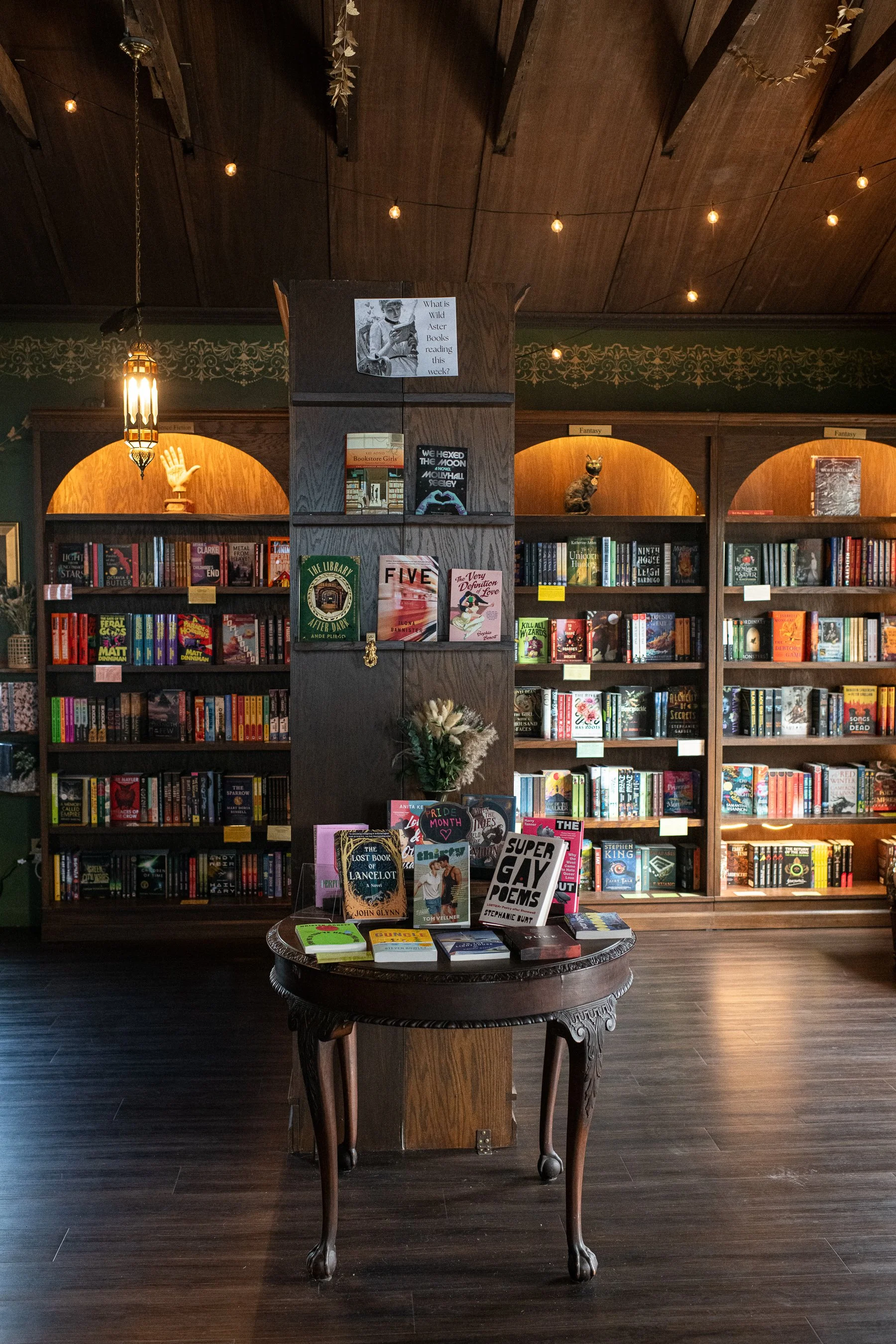 Indoor section of a bookstore with dark wooden floors and shelves filled with books. A round table in the foreground displays books, and there is a dark wooden column in the middle. Shelves behind the column feature decorative items like a ceramic ha