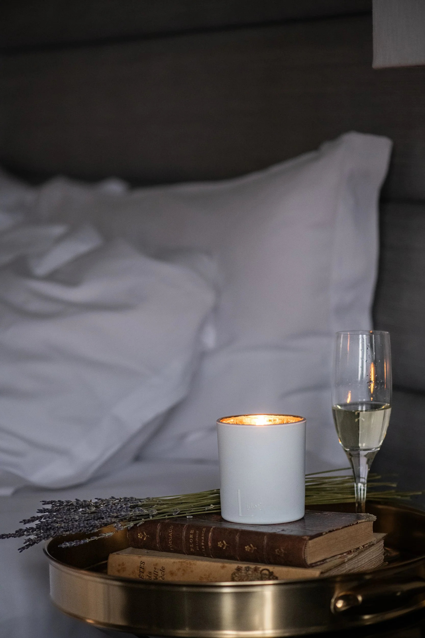 A tabletop with a lit white candle, a glass of white wine, a stack of vintage books, and dried lavender on a woven placemat against a backdrop of soft pillows and a candle