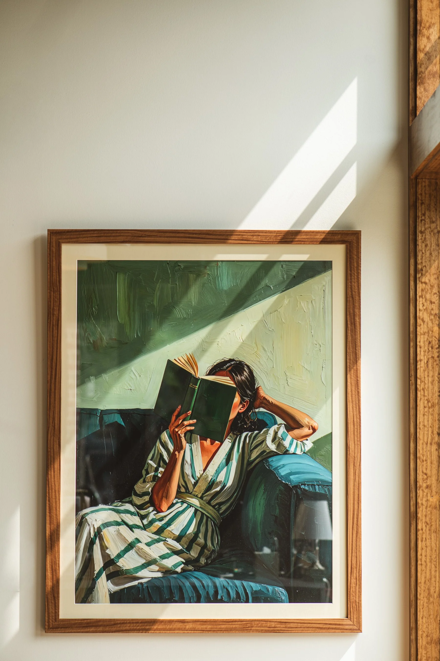 A painting of a woman with dark hair in a striped dress, sitting on a green chair, reading a book. The artwork is in a wooden frame and is displayed on a wall with sunlight casting shadows.
