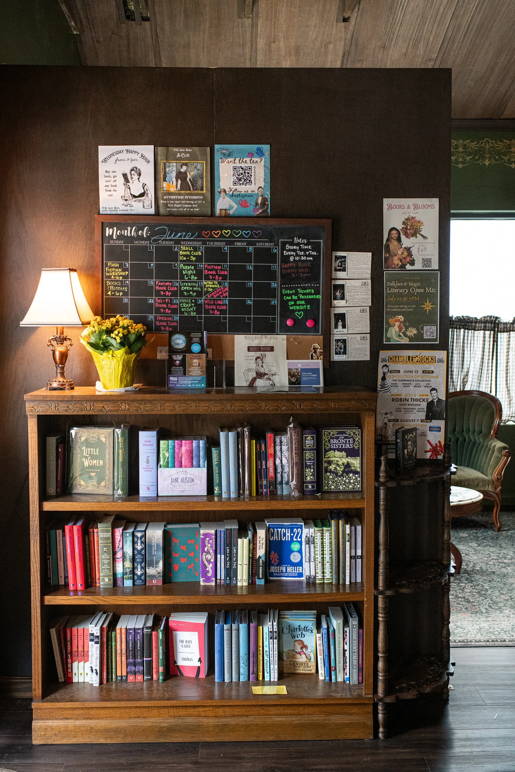A wooden bookshelf filled with various books, situated in a cozy room with a green armchair and a small side table. The top of the bookshelf has a decorative lamp with a yellow flower arrangement next to it. The bookshelf contains multiple shelves of