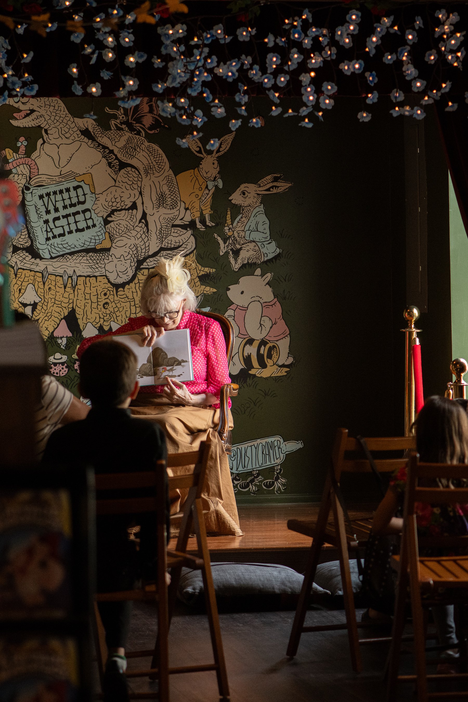 A woman with light-colored hair, glasses, and a red polka-dot shirt reading a picture book to children in a room decorated with a large book illustration and fairy lights.