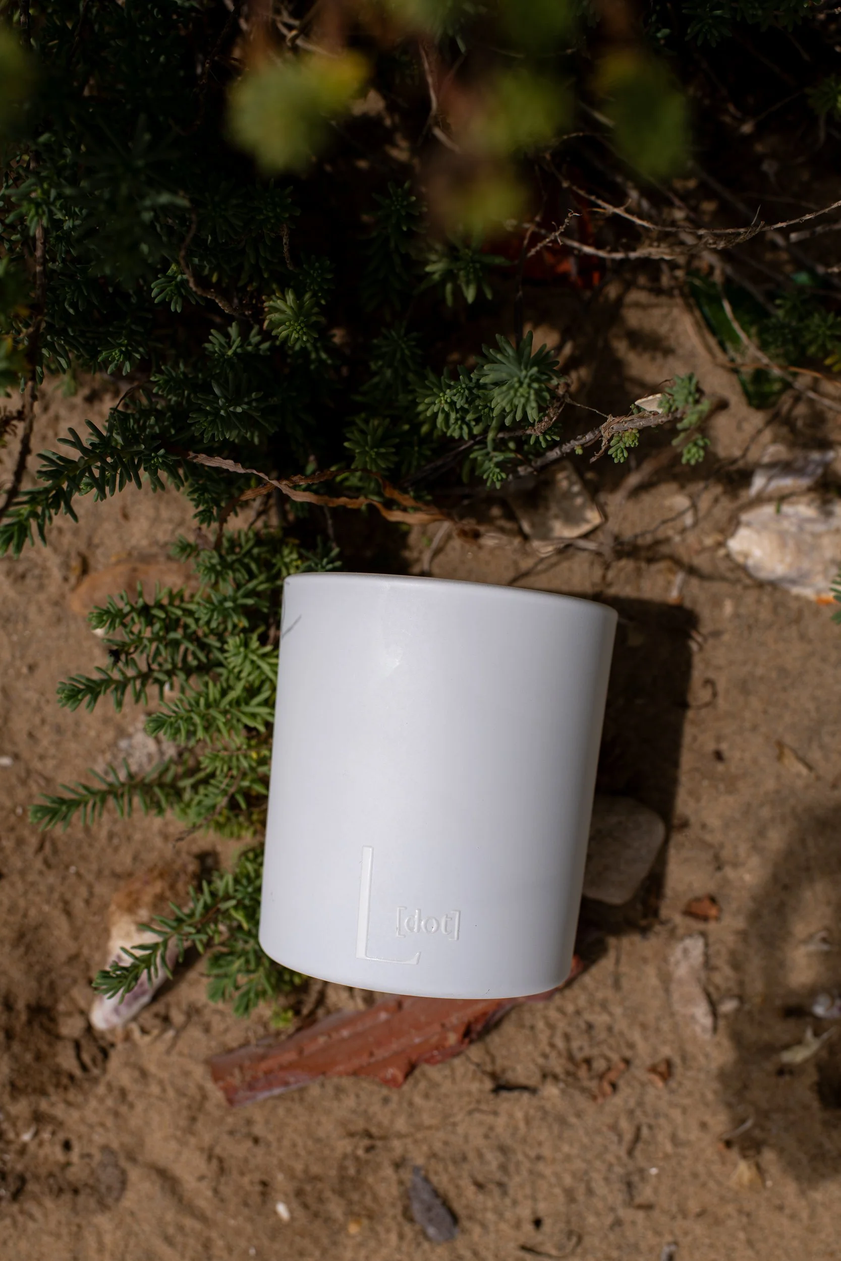 A white candle with rounded edges partially buried in dirt near green plants and small rocks.