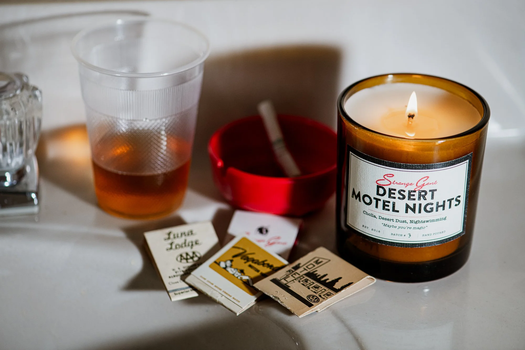 A lit candle labeled 'Desert Motel Nights,' a red ashtray with a cigarette, a plastic cup with a drink, and various matchbooks on a white surface.