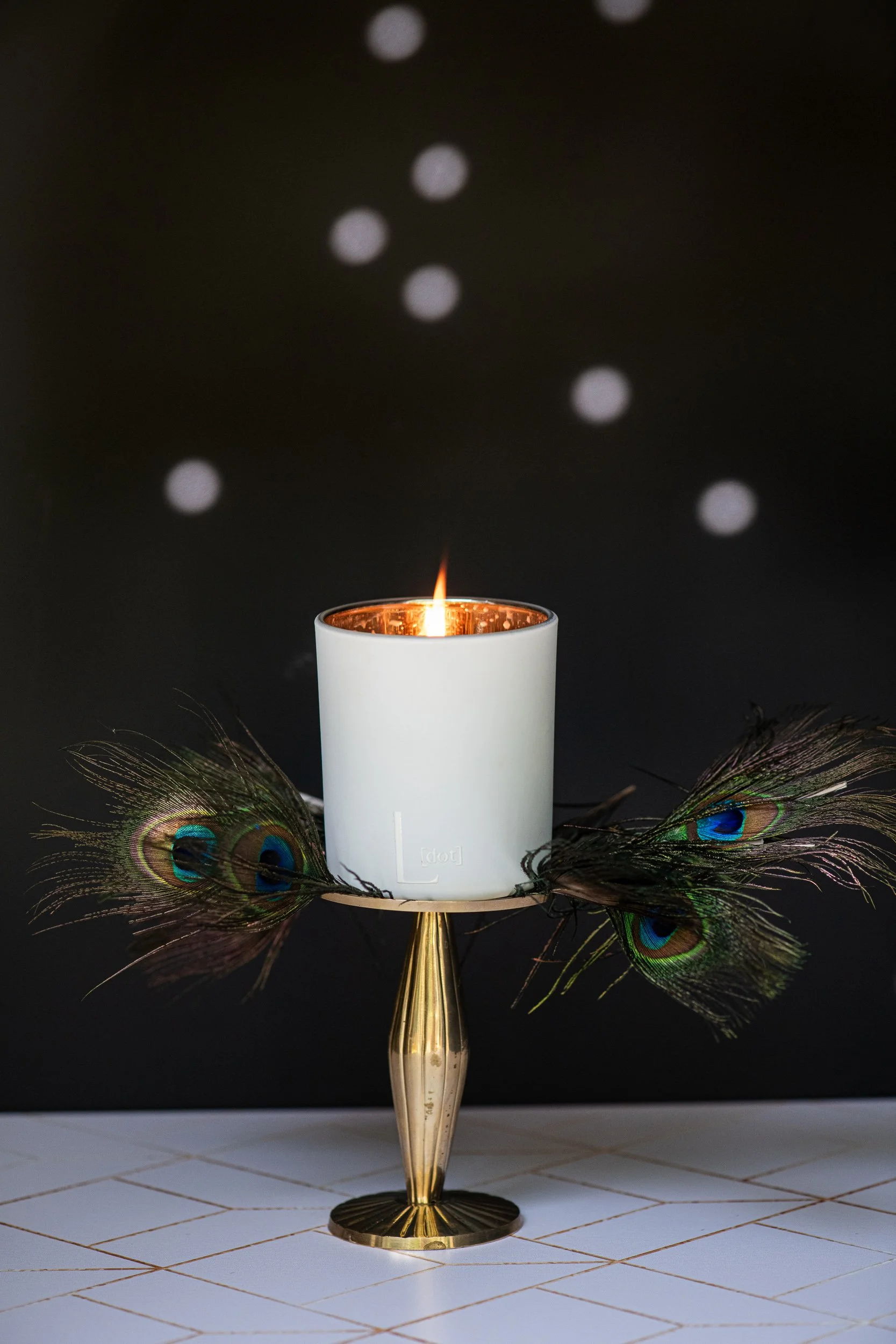 A white candle with a small flame, placed on a decorative stand, surrounded by peacock feathers on a tiled surface, against a black background with bokeh light effects.