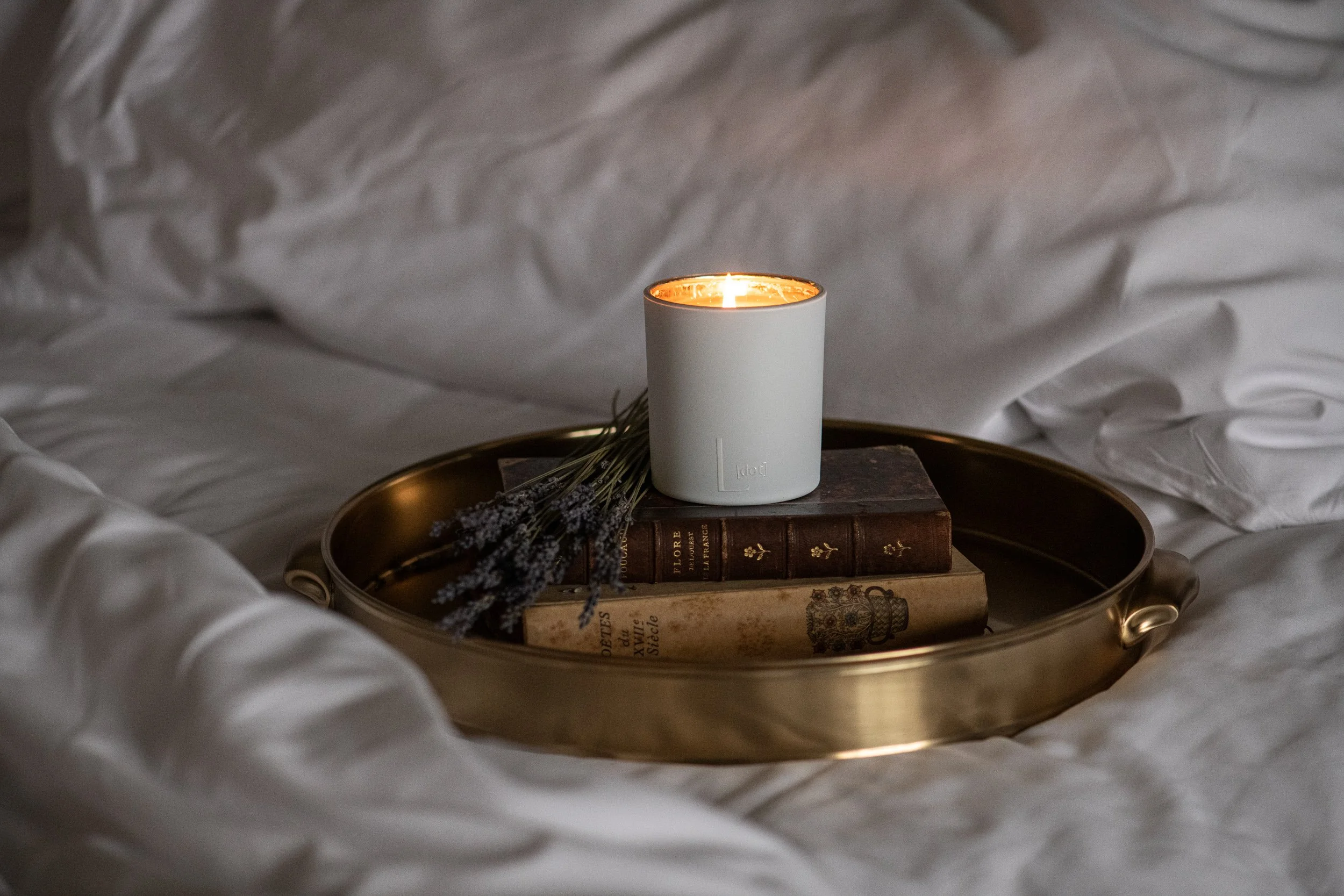 A white candle burning on top of vintage books, placed on a round metallic tray amid crumpled white bedsheets, with dried lavender bunches nearby.