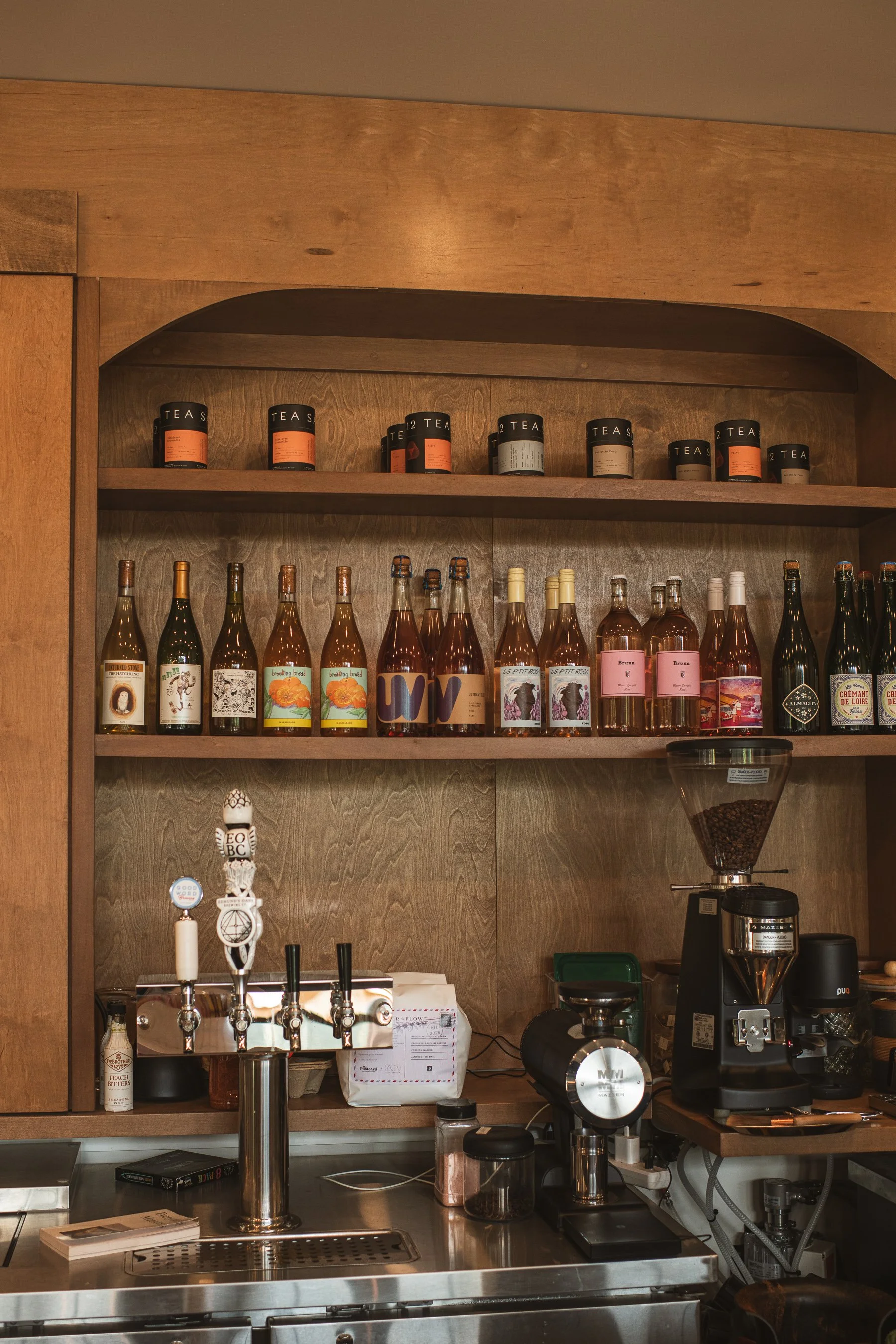 Wooden shelves with assorted tea containers and bottles, coffee bar with coffee machine and grinder, coffee beans, and various bar tools.