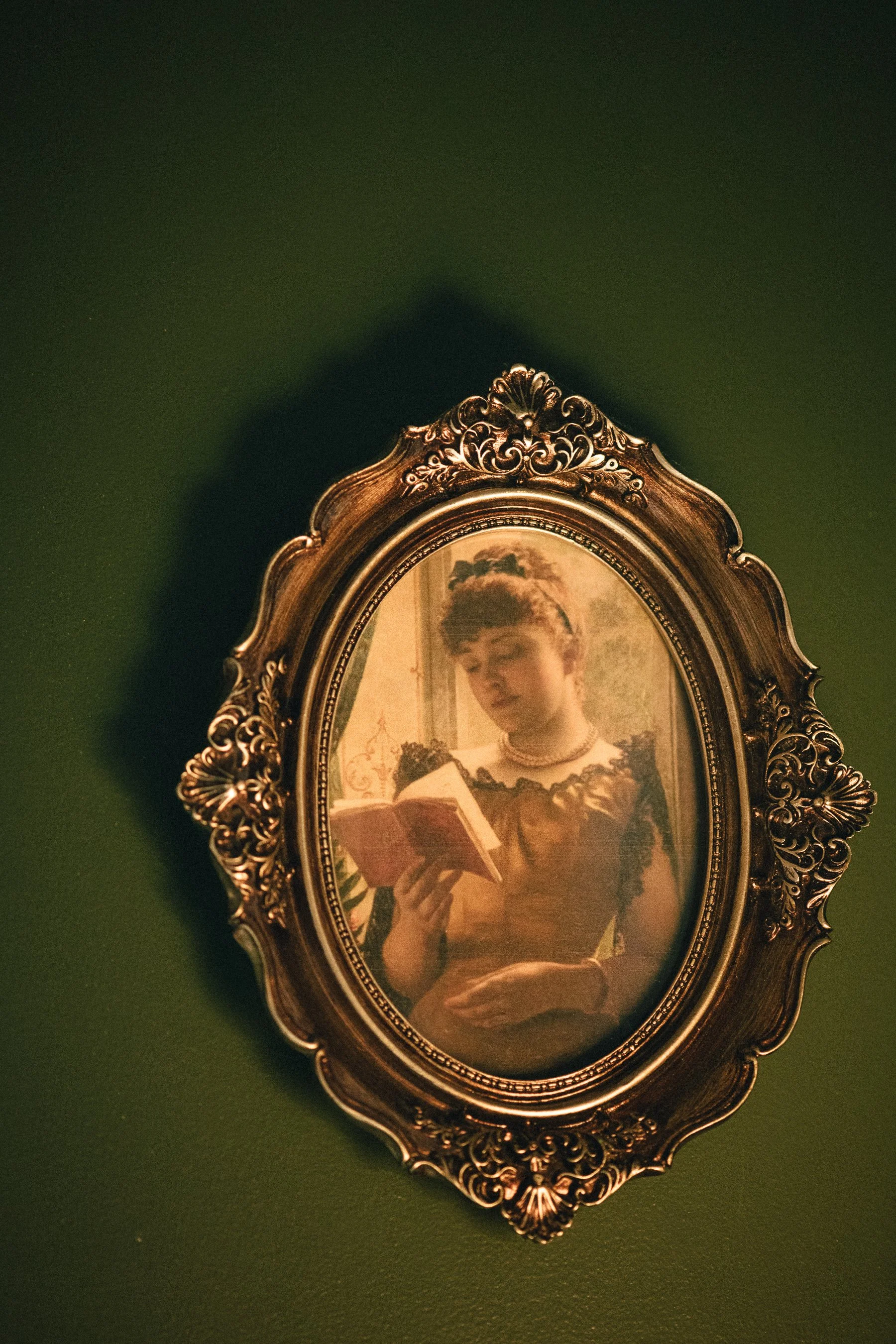 An ornate gold-framed oval mirror on a green wall. The mirror reflects a woman with short hair reading a book.