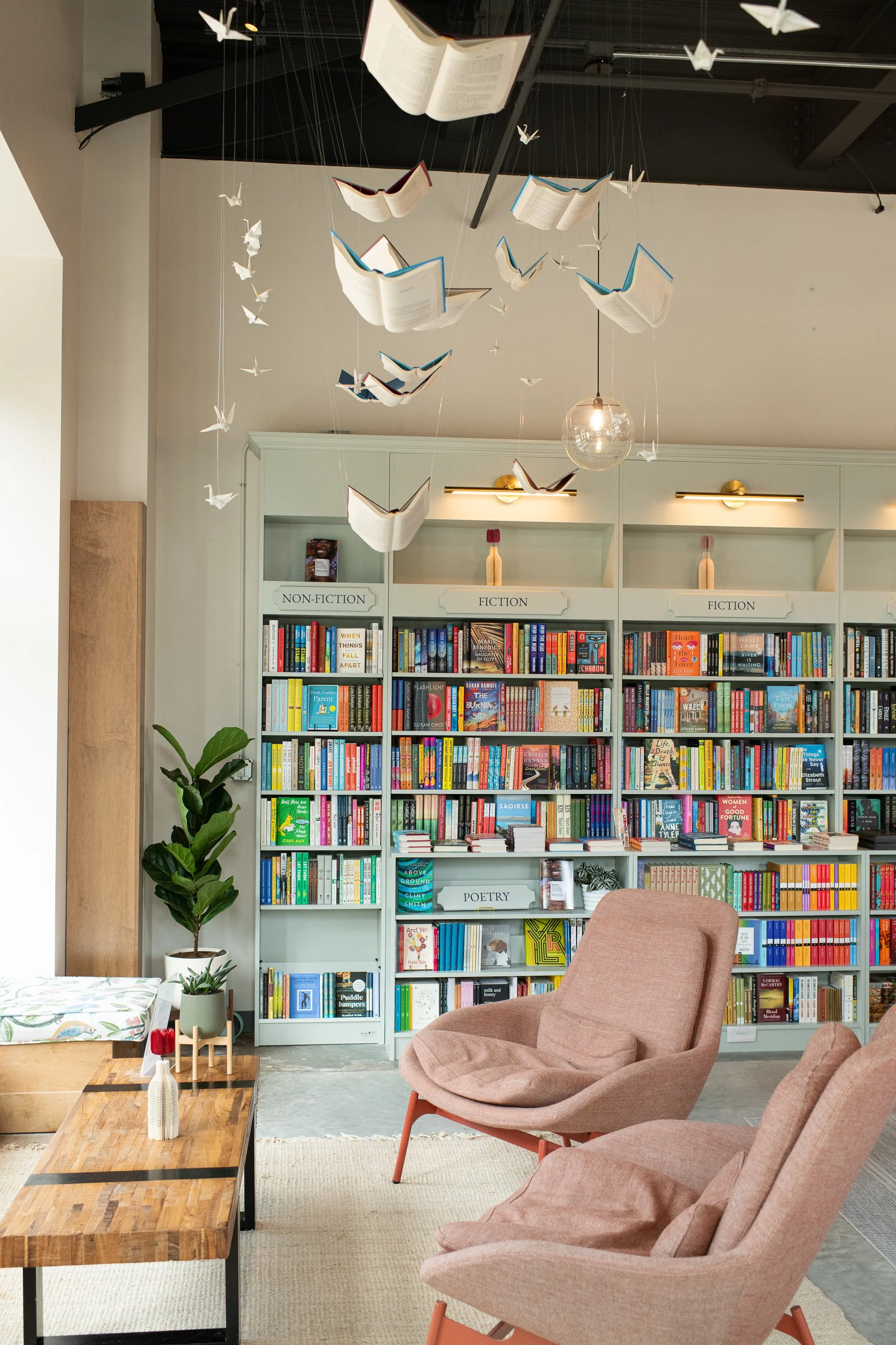 A cozy bookstore interior with beige and white bookshelves labeled 'Non-Fiction' and 'Poetry', filled with colorful books. Hanging from the ceiling are open book and origami crane decorations. To the left, a potted plant sits on the floor. In the for