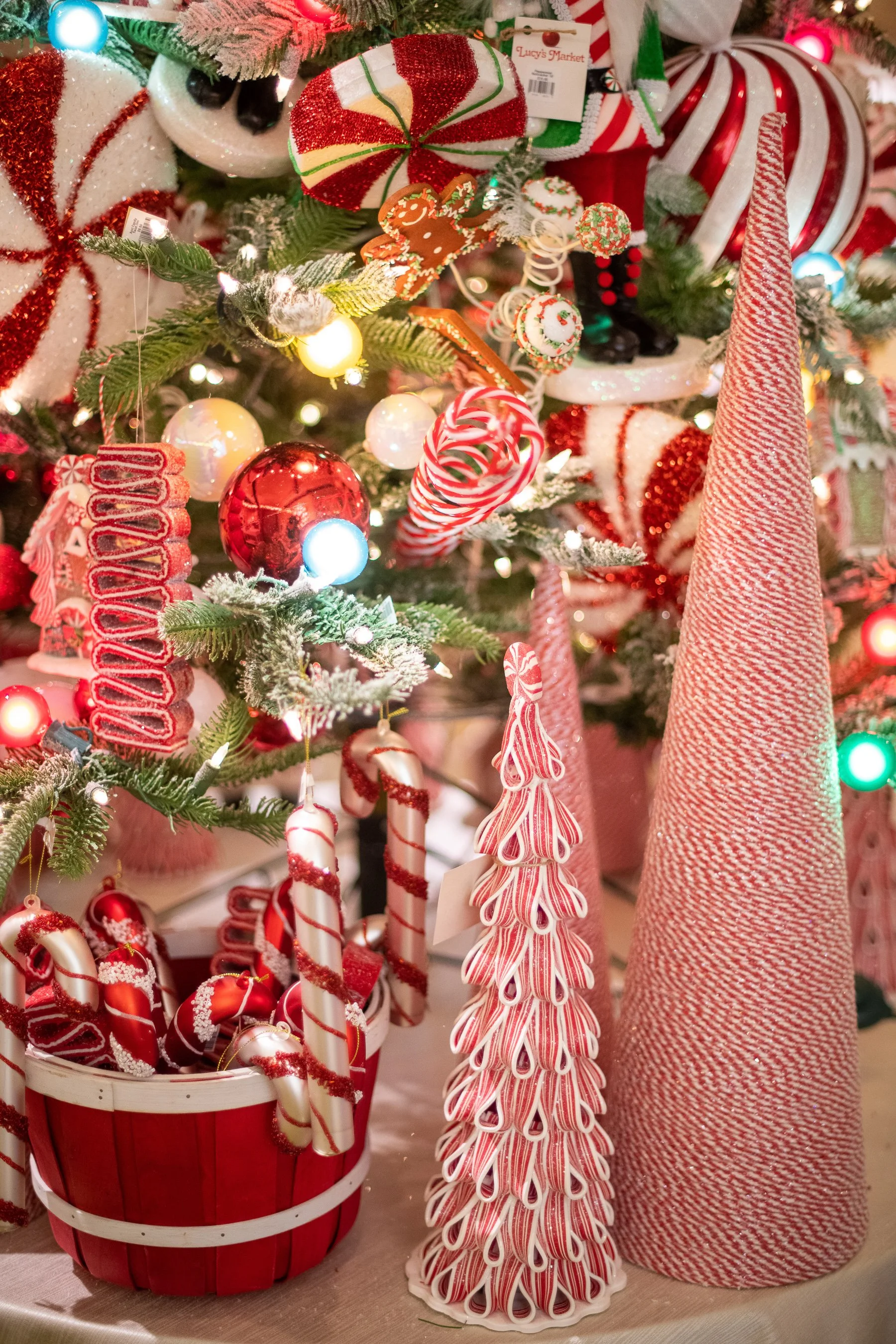 A decorated Christmas tree with various red, white, and green ornaments, including candy canes, a gingerbread man, and a striped tree-shaped decoration, with a red basket filled with candy canes and a red and white cone-shaped ornament nearby.