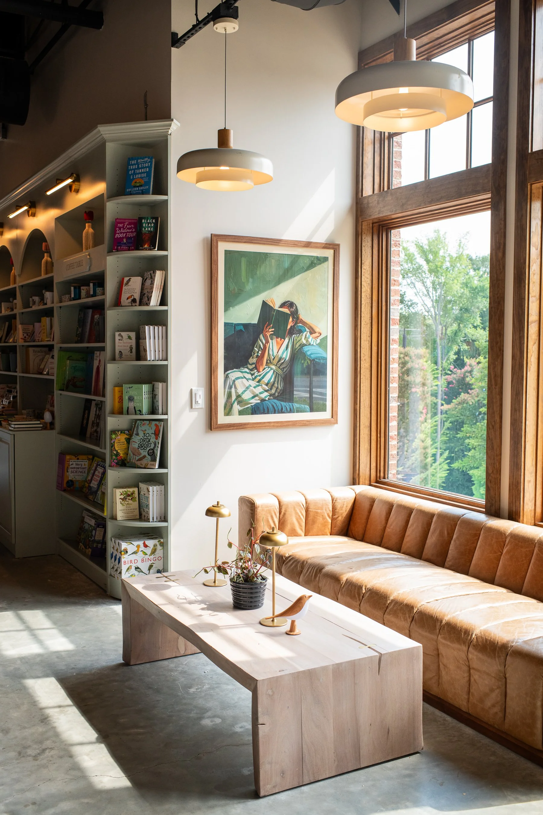 Interior of a cozy coffee shop or cafe with a large window, a tan velvet sofa, a framed artwork of a woman reading a book, a light wooden coffee table with small decorative lamps and a plant, a white bookshelf filled with books, and modern hanging pe