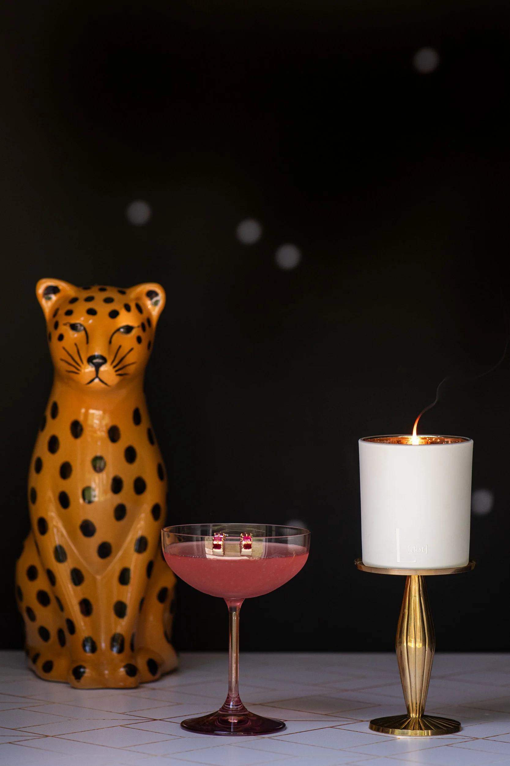 A ceramic cheetah statue, a pink cocktail glass with rings, and a white candle on a table against a black background.