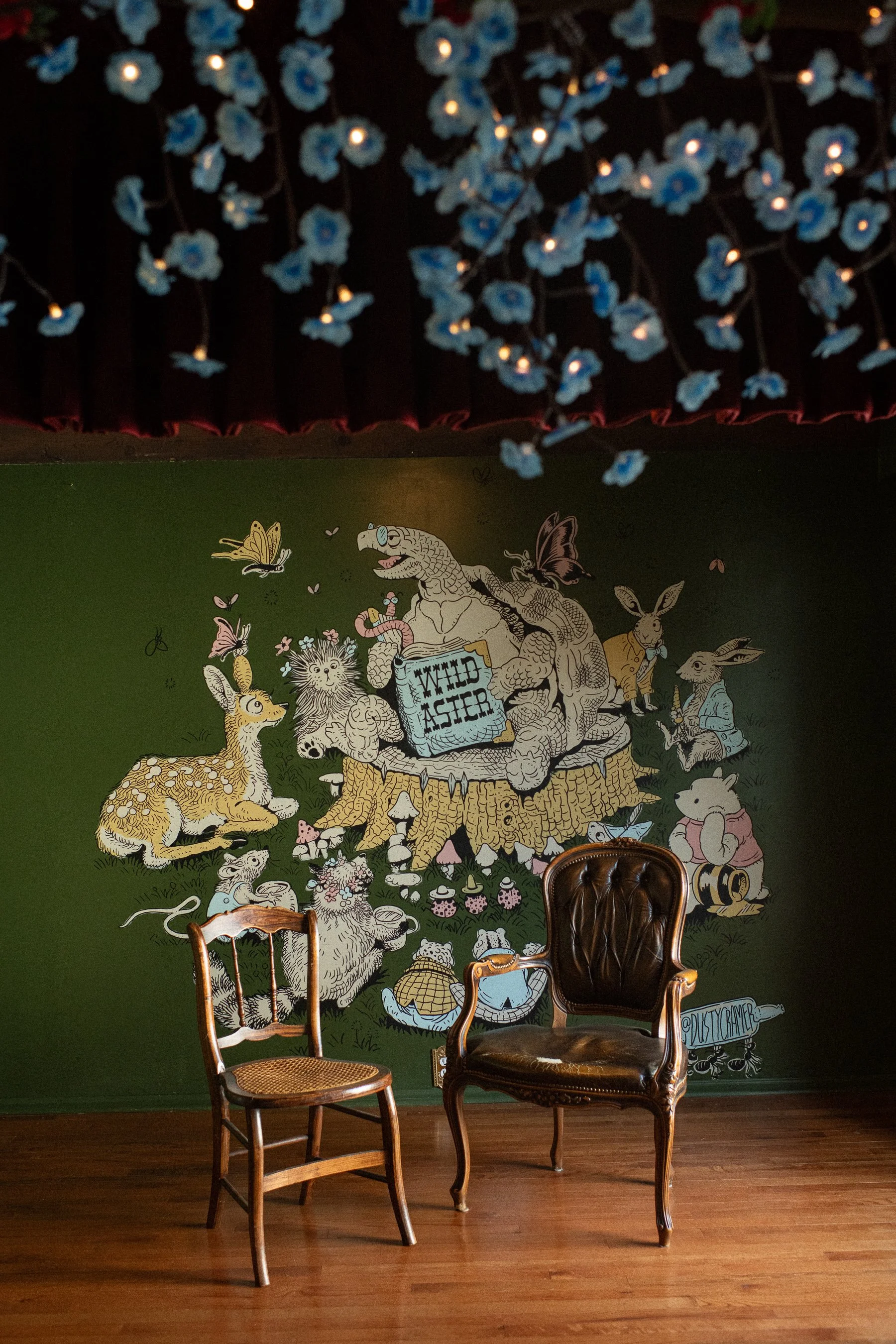 Indoor scene with a colorful mural of animals and mythical creatures on a green wall, a wooden chair with a cane seat, and a vintage leather armchair with curved wood armrests and legs.
