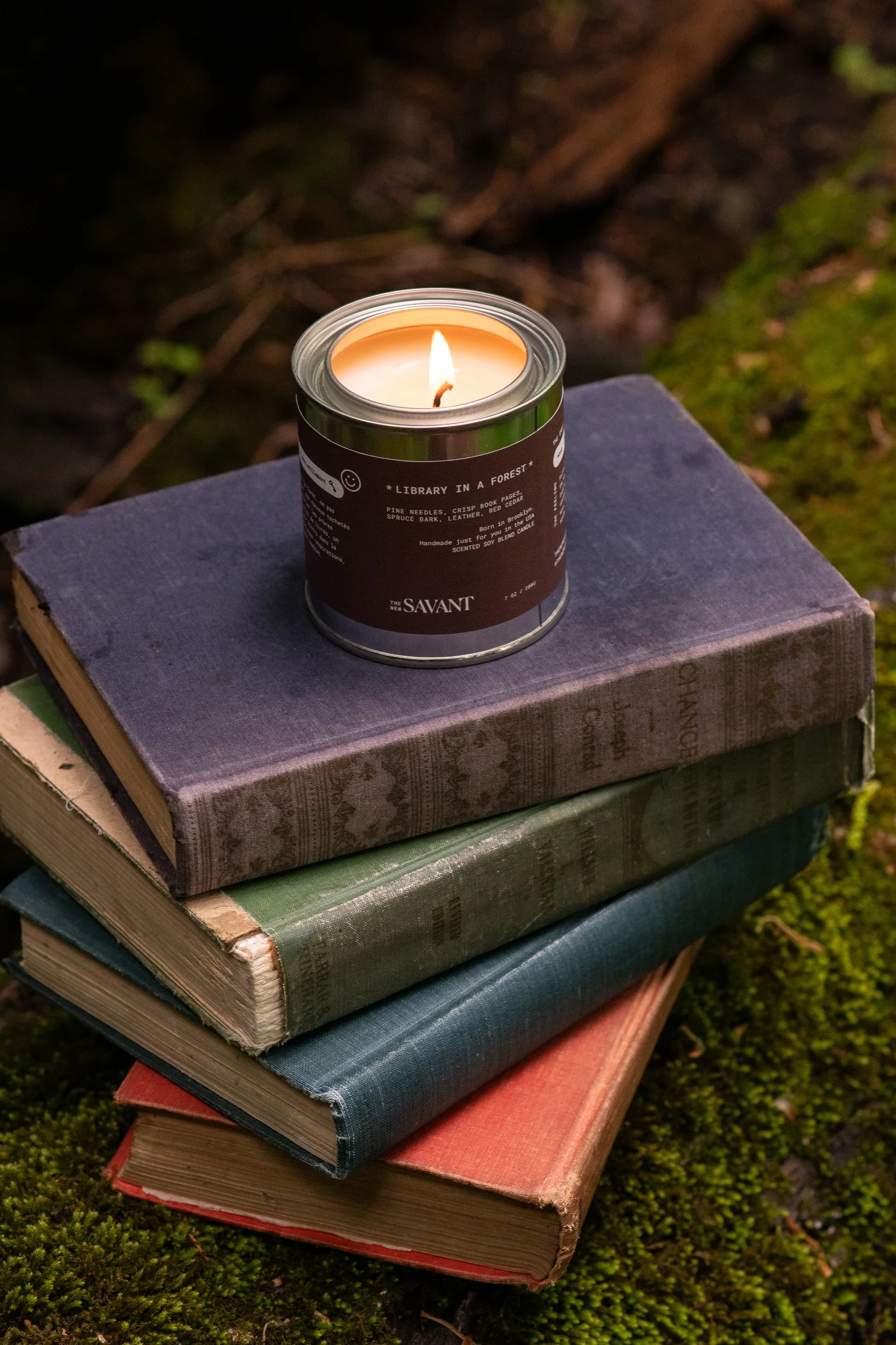 Lit Library in a Forest candle resting on a stack of vintage books surrounded by moss and woodland textures.