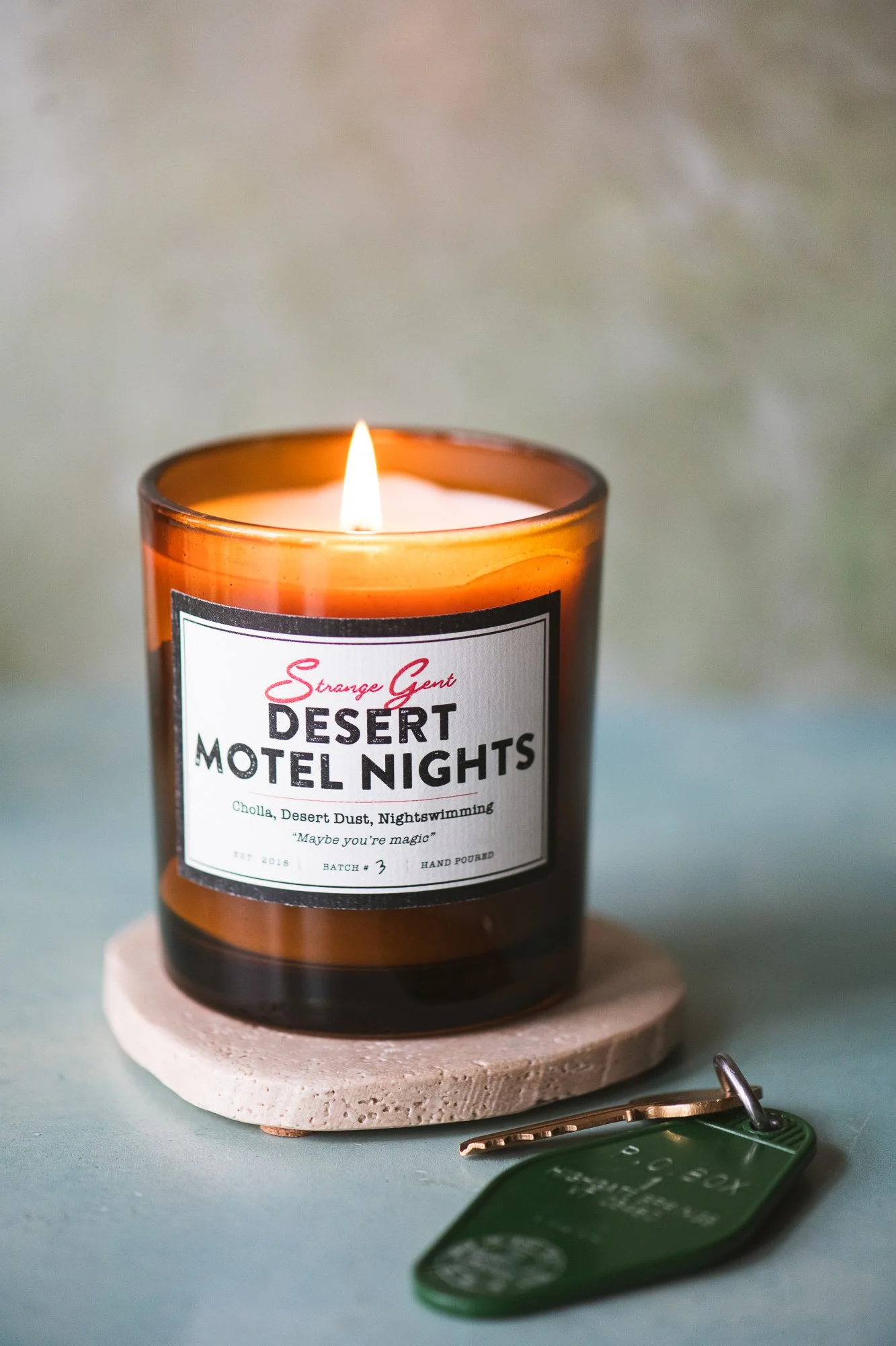 A lit amber glass candle labeled "Desert Motel Nights" on a white coaster, with a green key fob nearby, on a light blue surface.