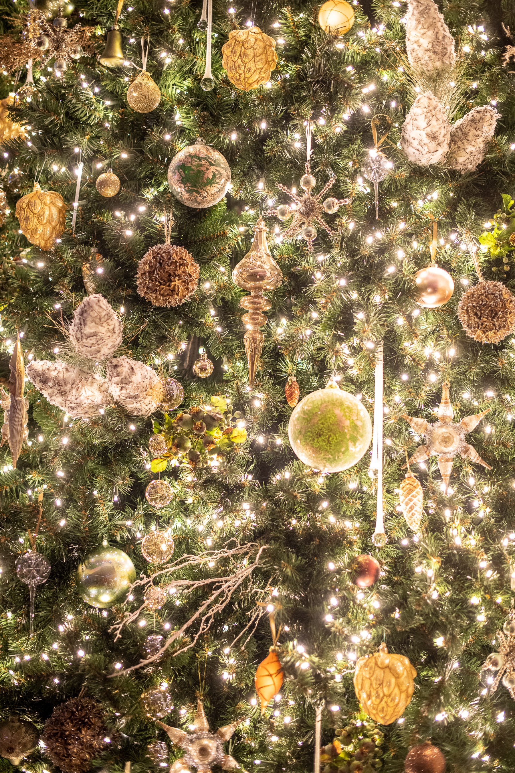A decorated Christmas tree with gold, silver, and clear ornaments, pinecones, and white lights.