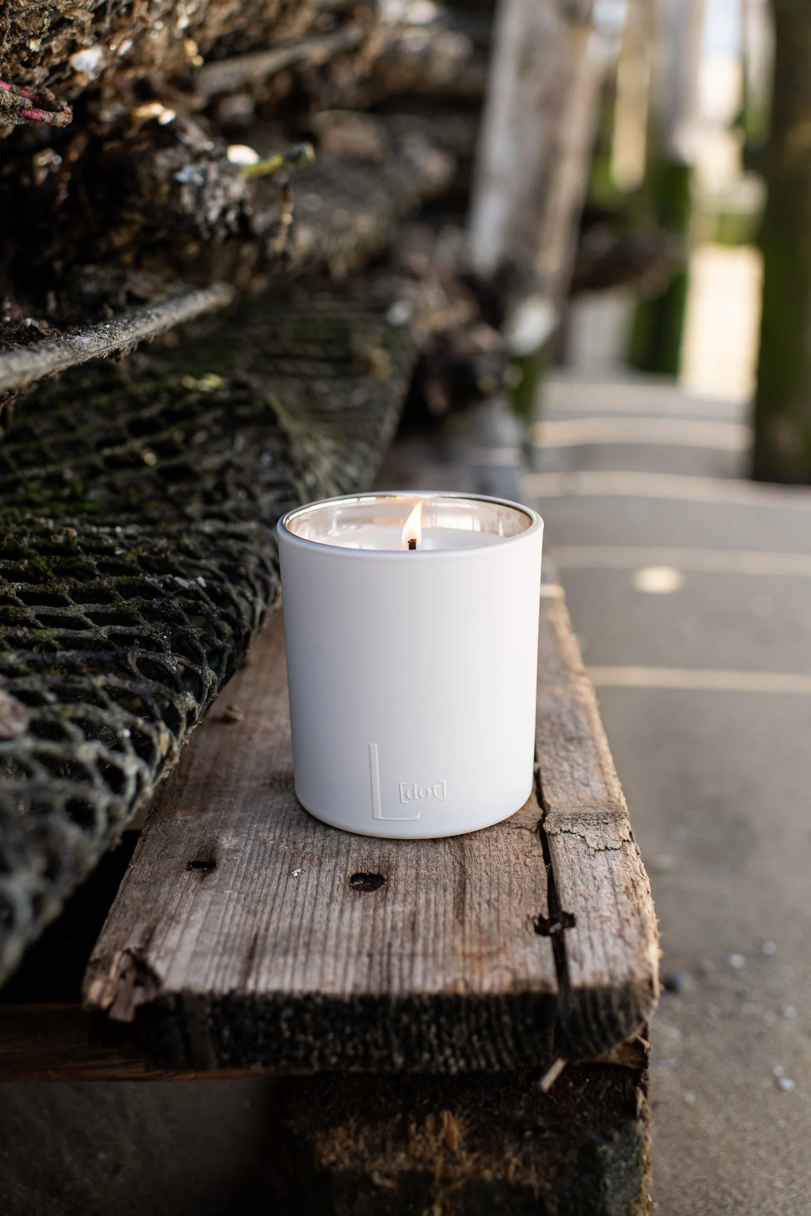 A white candle with a flame on a weathered wooden surface outdoors, with a rustic wooden fence and blurred background.