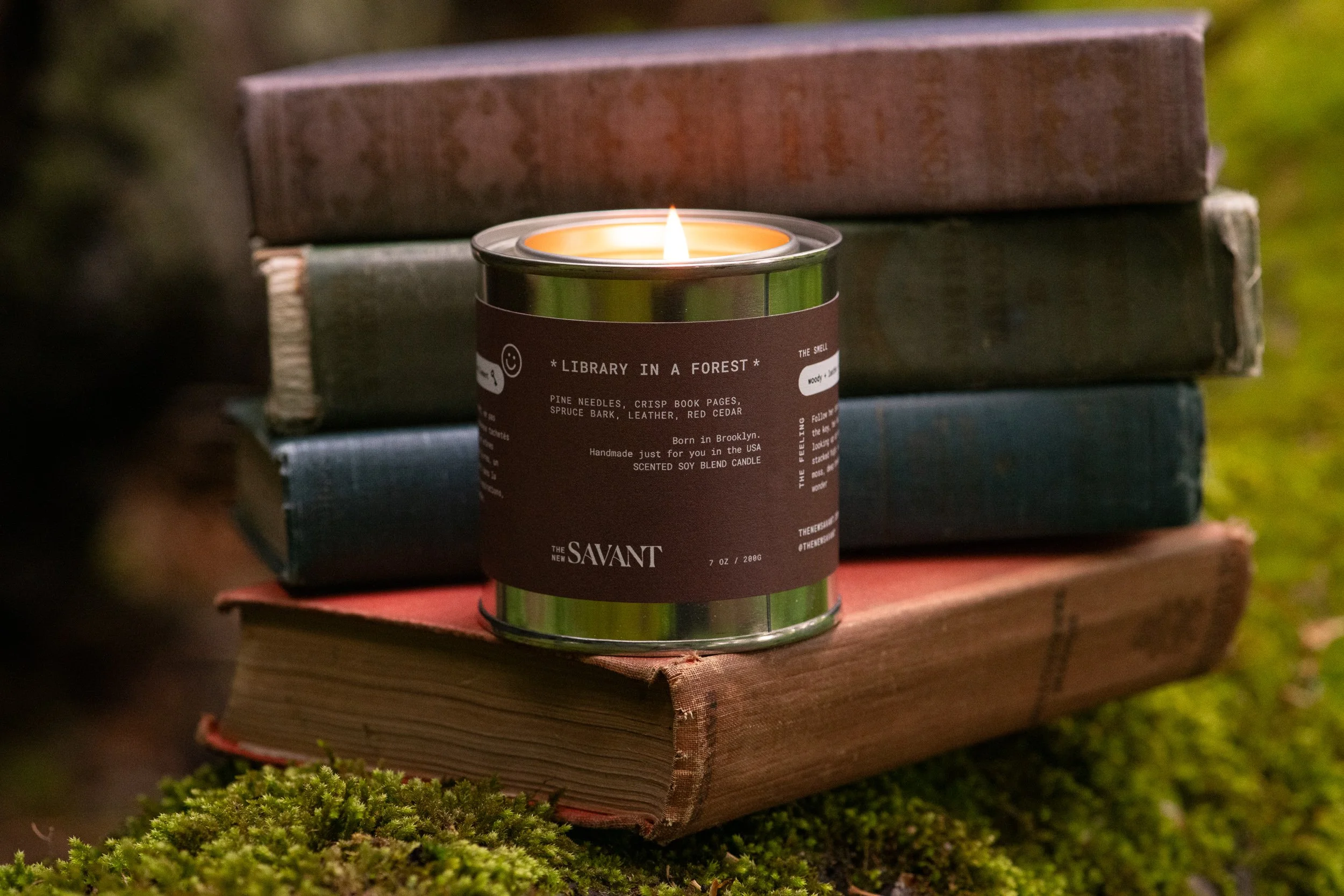 A lit candle in a metal container sits on a stack of three worn books outdoors on a mossy surface, with a blurred grassy background.