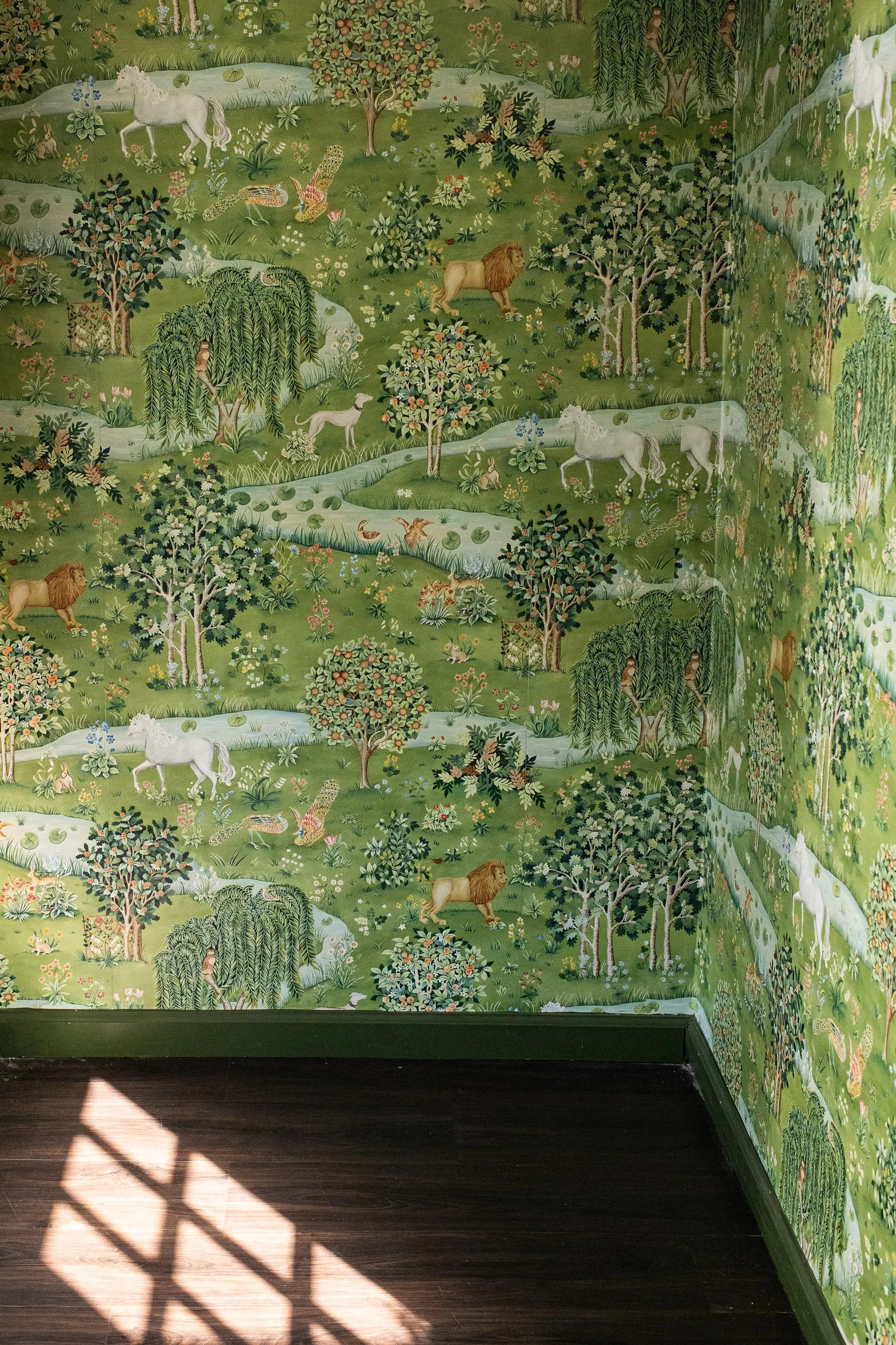 Decorative wallpaper at indpendent bookstore Wild Aster in Chamblee, GA with pastoral scene of lions, horses, deer, rabbits, birds, and trees in a lush, green landscape on the walls of a room with dark wooden flooring.