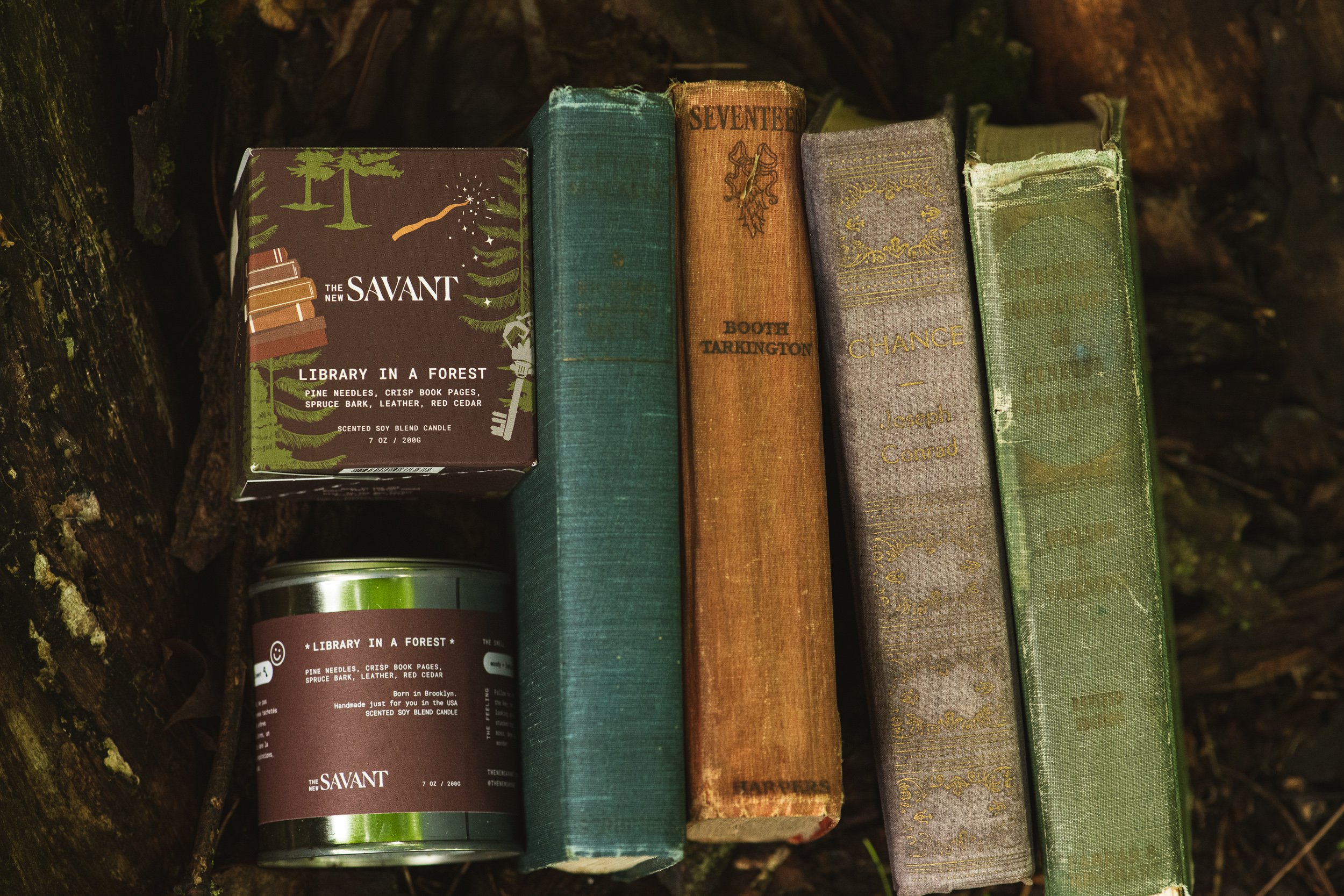 A row of vintage hardcover books outdoors, next to a candles in small tins with forest-themed designs and text.