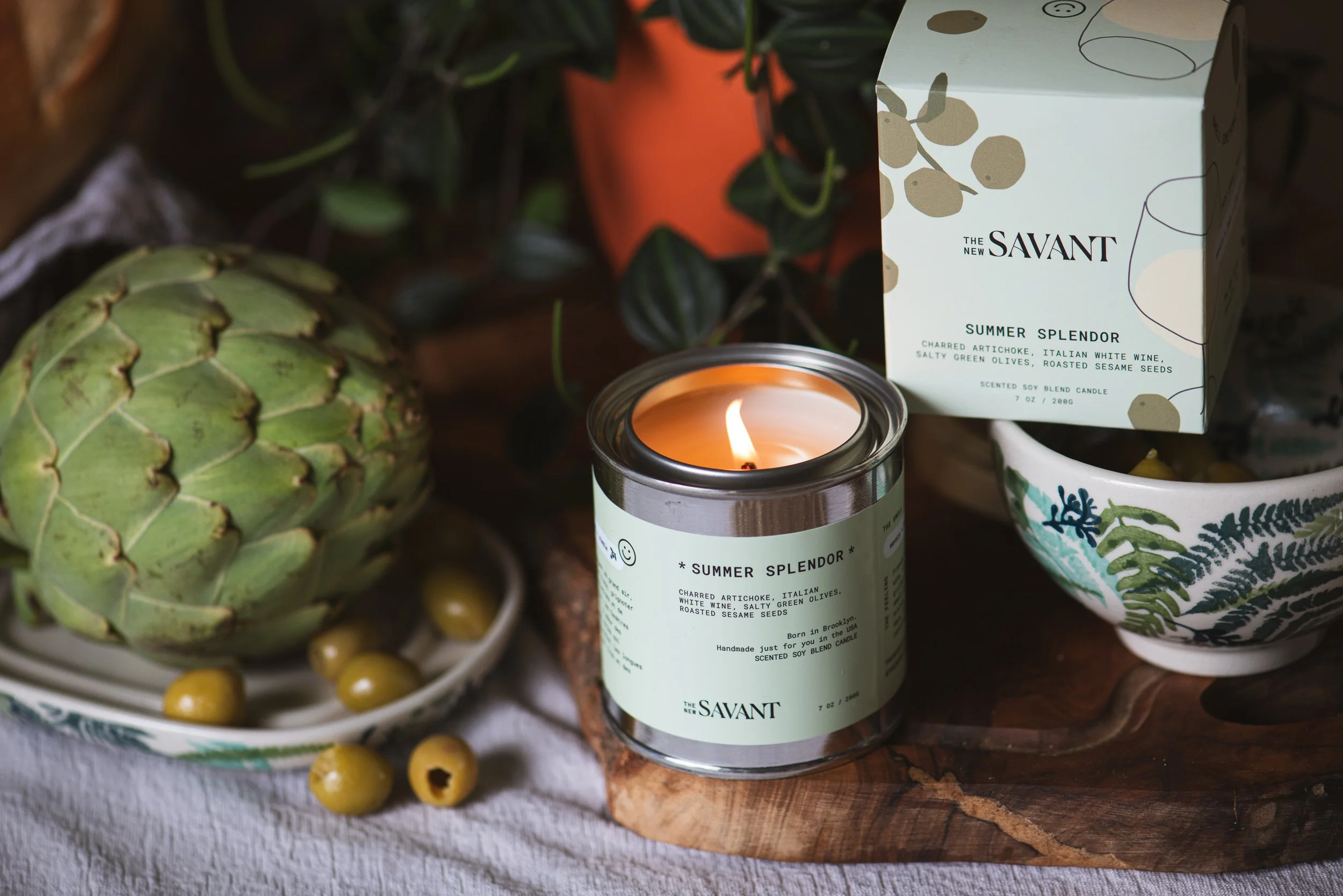 A candle labeled "Summer Splendor" surrounded by a pineapple, green olives, and a bowl with a decorative leaf pattern.
