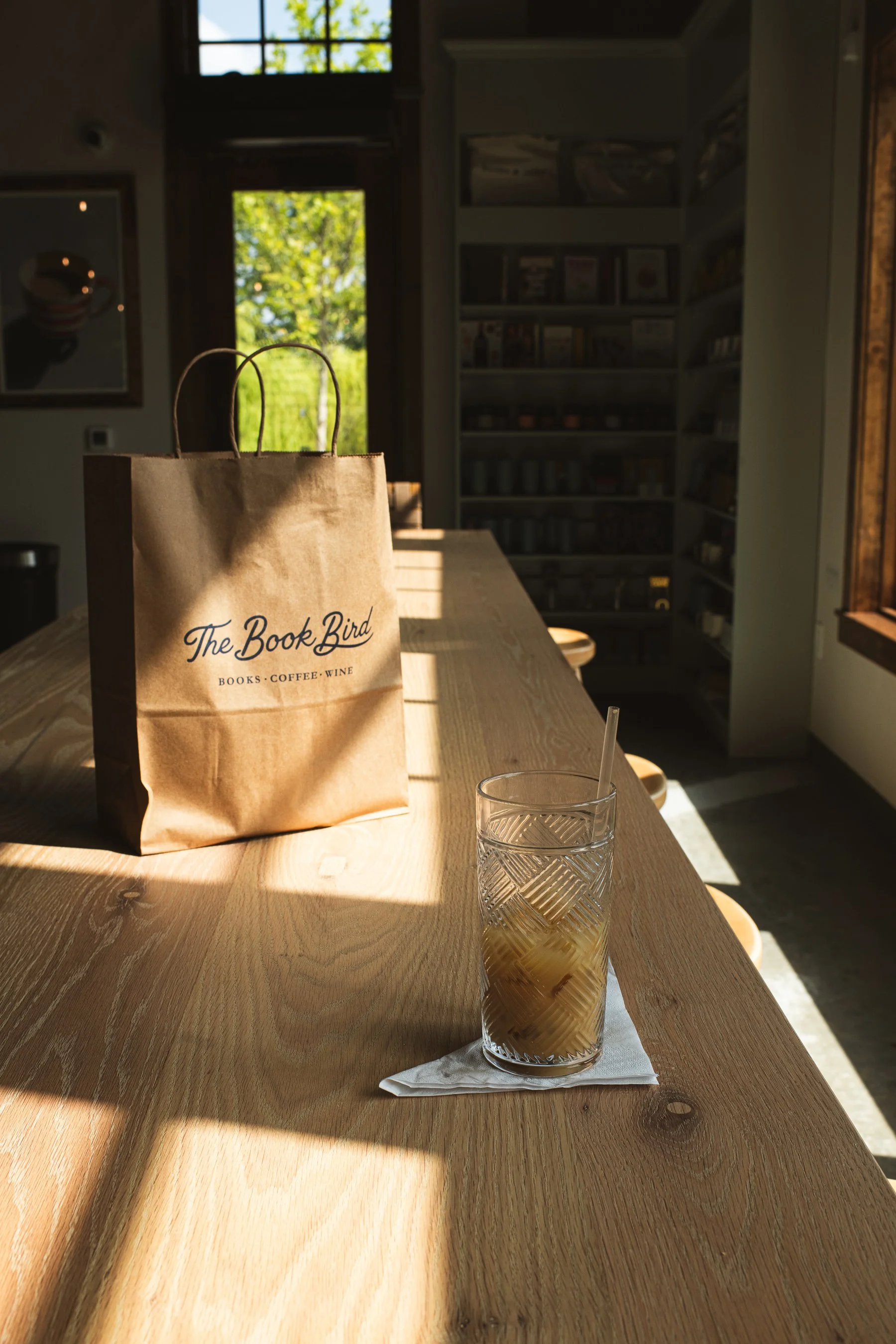 A paper bag labeled "The Book Bird" on a wooden table, with a glass of iced tea and a paper napkin in front of it, in a cozy cafe with bookshelves and windows letting in natural light.