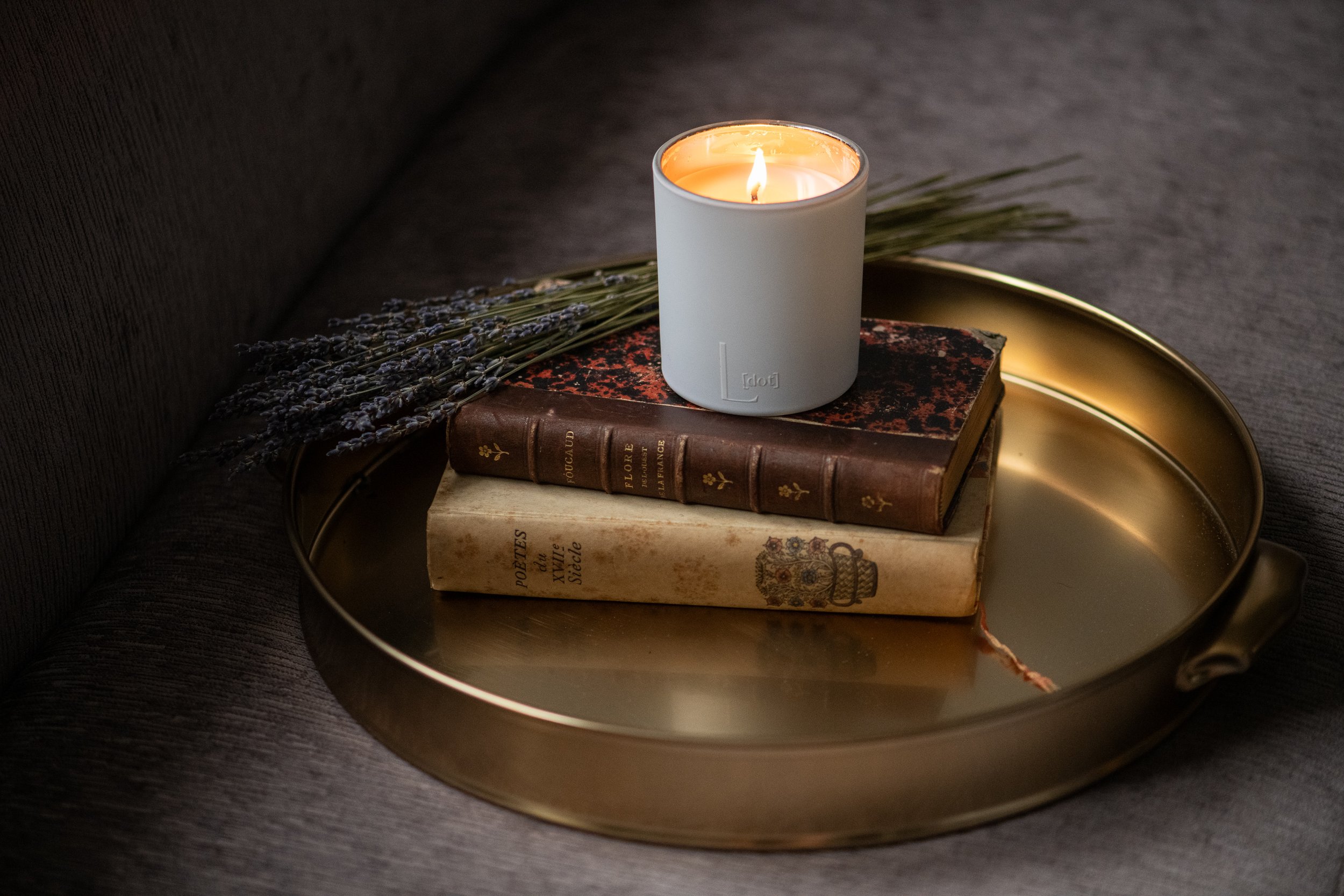A lit white candle on top of three stacked vintage books, with a bunch of dried lavender on a gold tray.