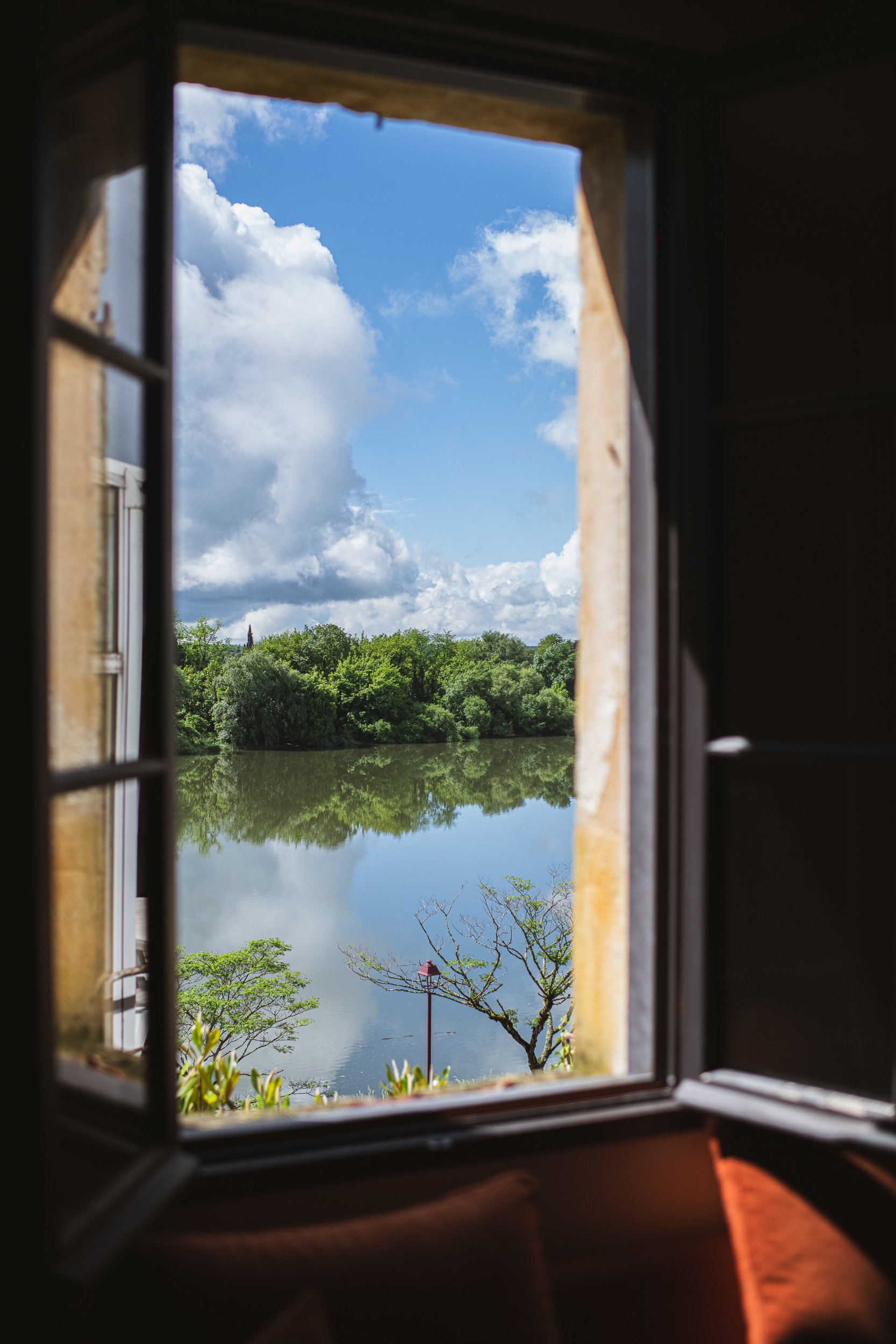 View of a lake with green trees, cloudy sky, and a lamp post seen through a square window with a wooden frame.
