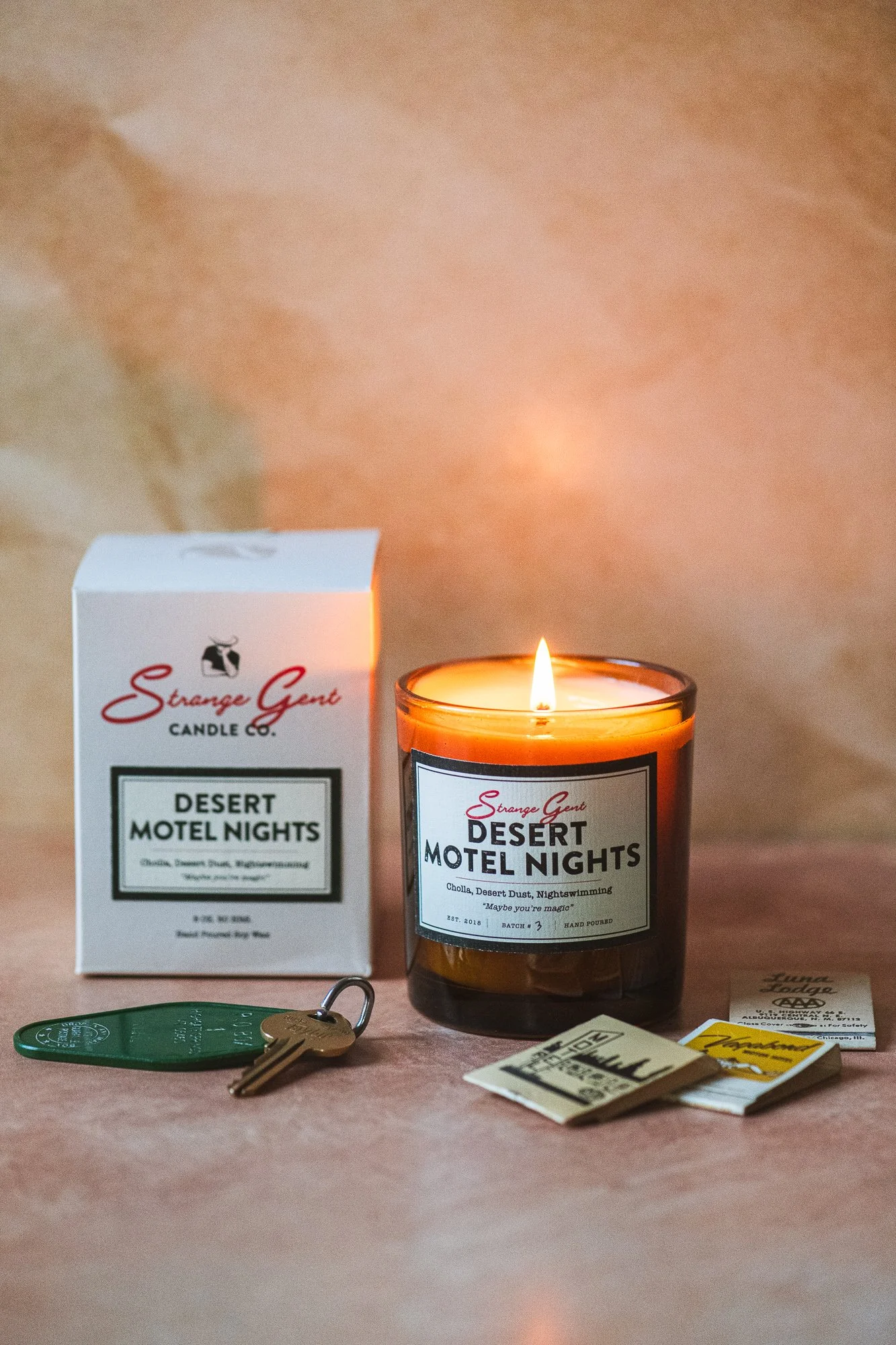 A lit candle in a brown glass jar labeled 'Strange Gentry Desert Motel Nights' on a wooden surface, with a white box of the same candle, a key with a green keychain, and some paper receipts or notes nearby.