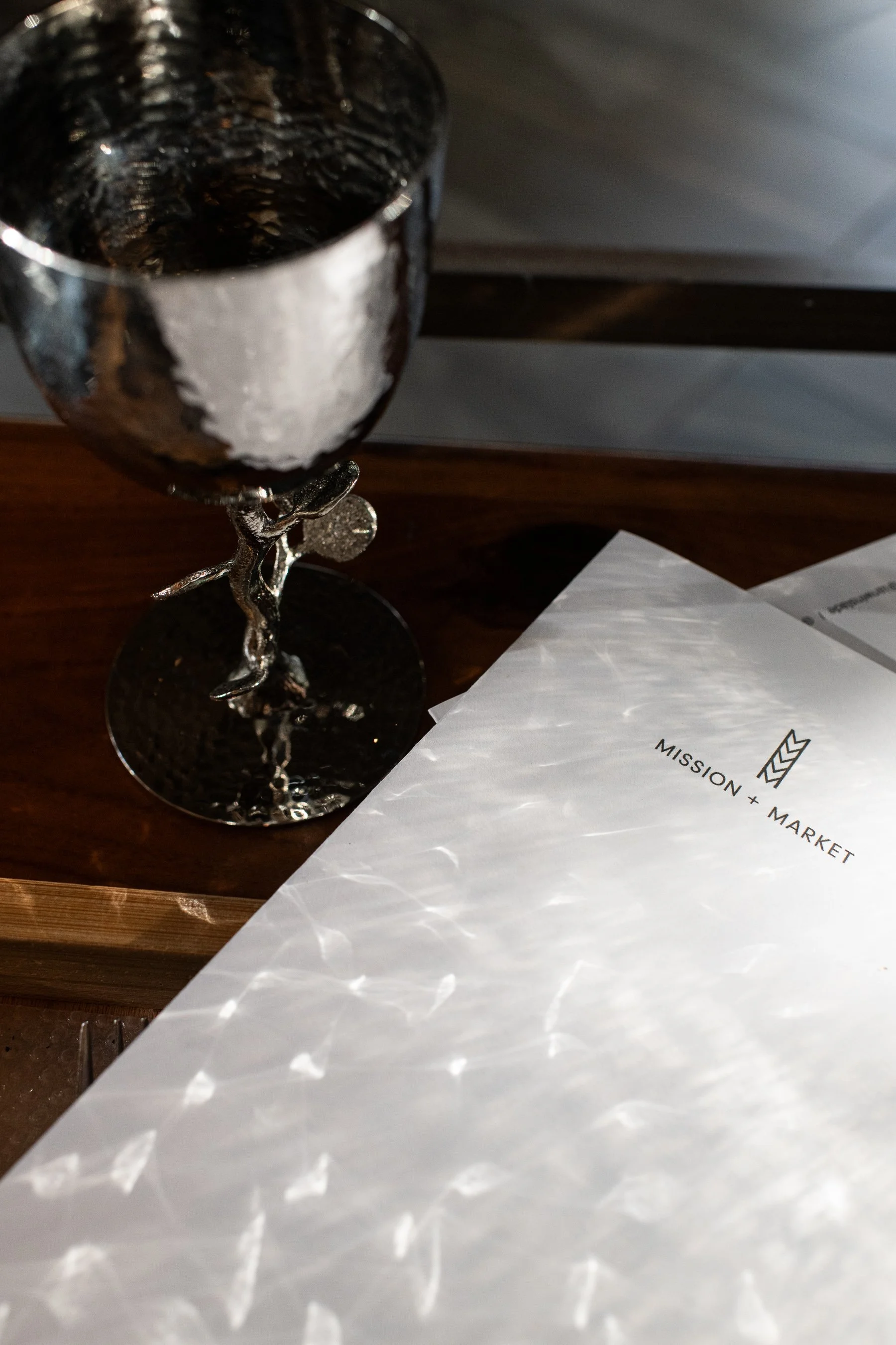 A silver trophy with a figure holding a shield near the top, placed on a wooden surface next to a piece of paper with text. The trophy reflects light, creating a pattern of reflections on the paper.