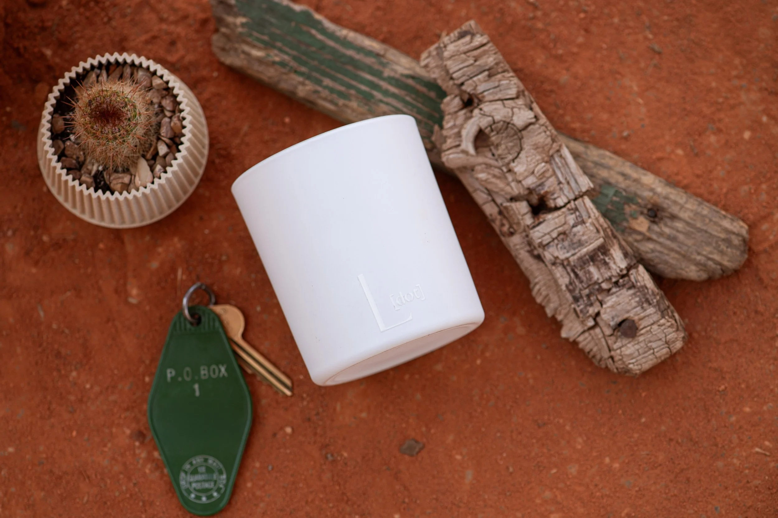 A small round cactus in a beige ribbed pot with rocks, a white candle, a weathered green and brown wooden stick, a green keychain labeled 'P.O. BOX 1', and a wooden stick with chipped paint on a reddish-brown dirt surface.