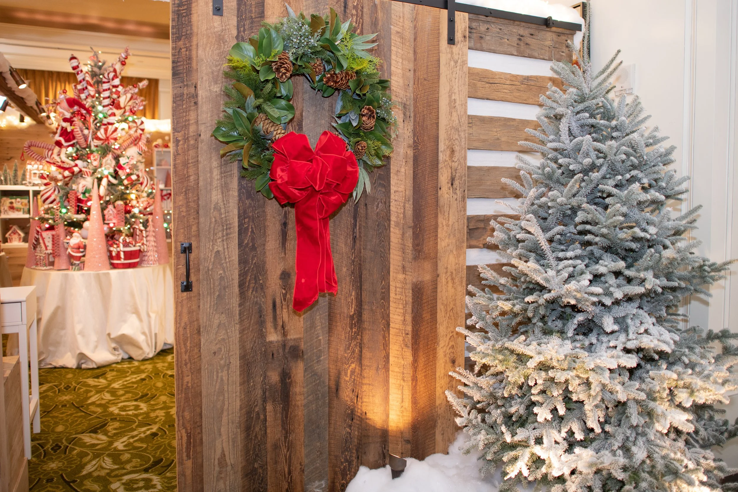 A Christmas-themed display featuring a snowy evergreen tree beside a wooden divider, with a holiday wreath decorated with pinecones, green leaves, and a red velvet bow hanging on the divider.