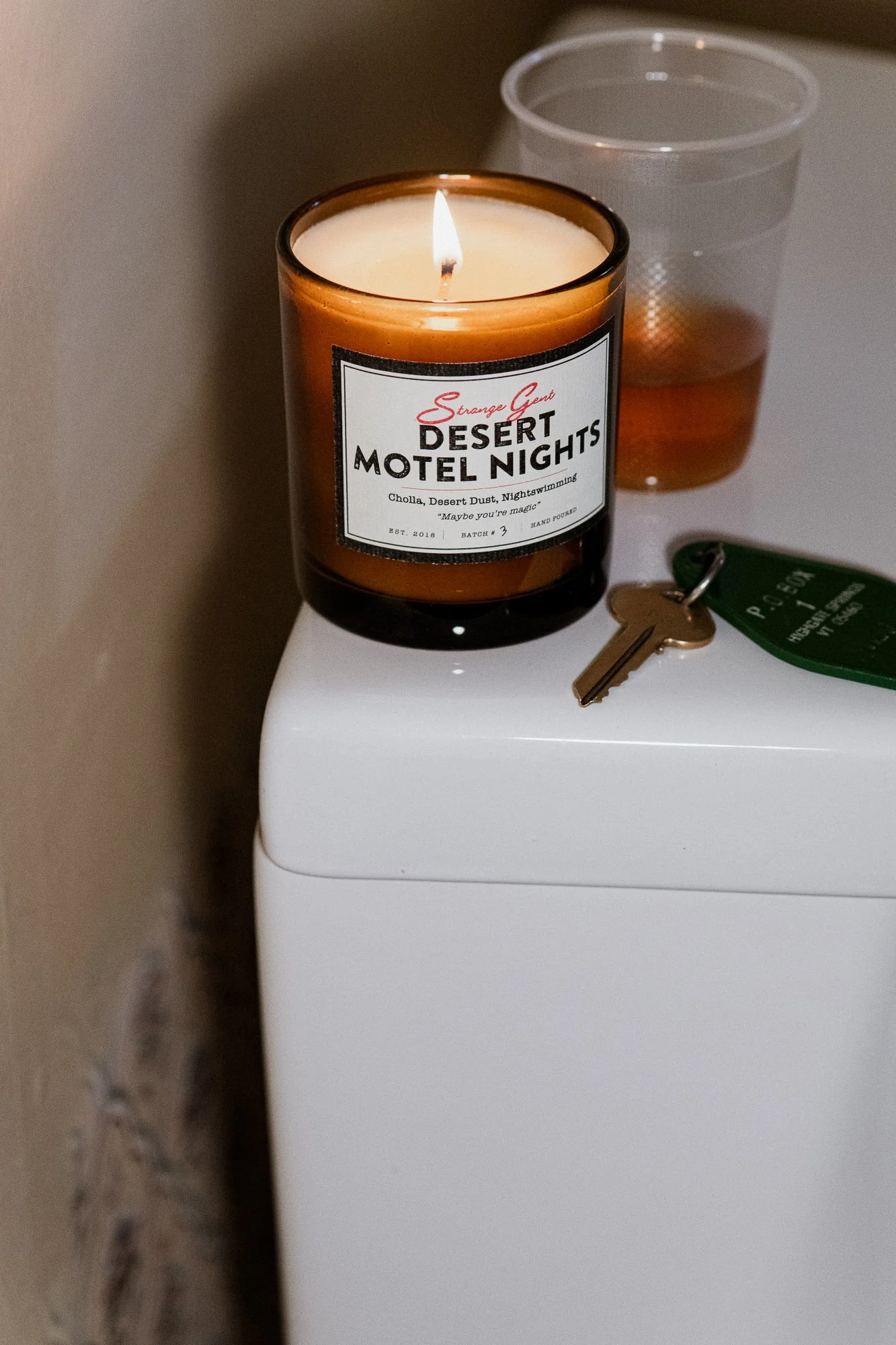 A lit candle labeled 'Desert Motel Nights' placed on top of a white surface with a glass of water and a key nearby.