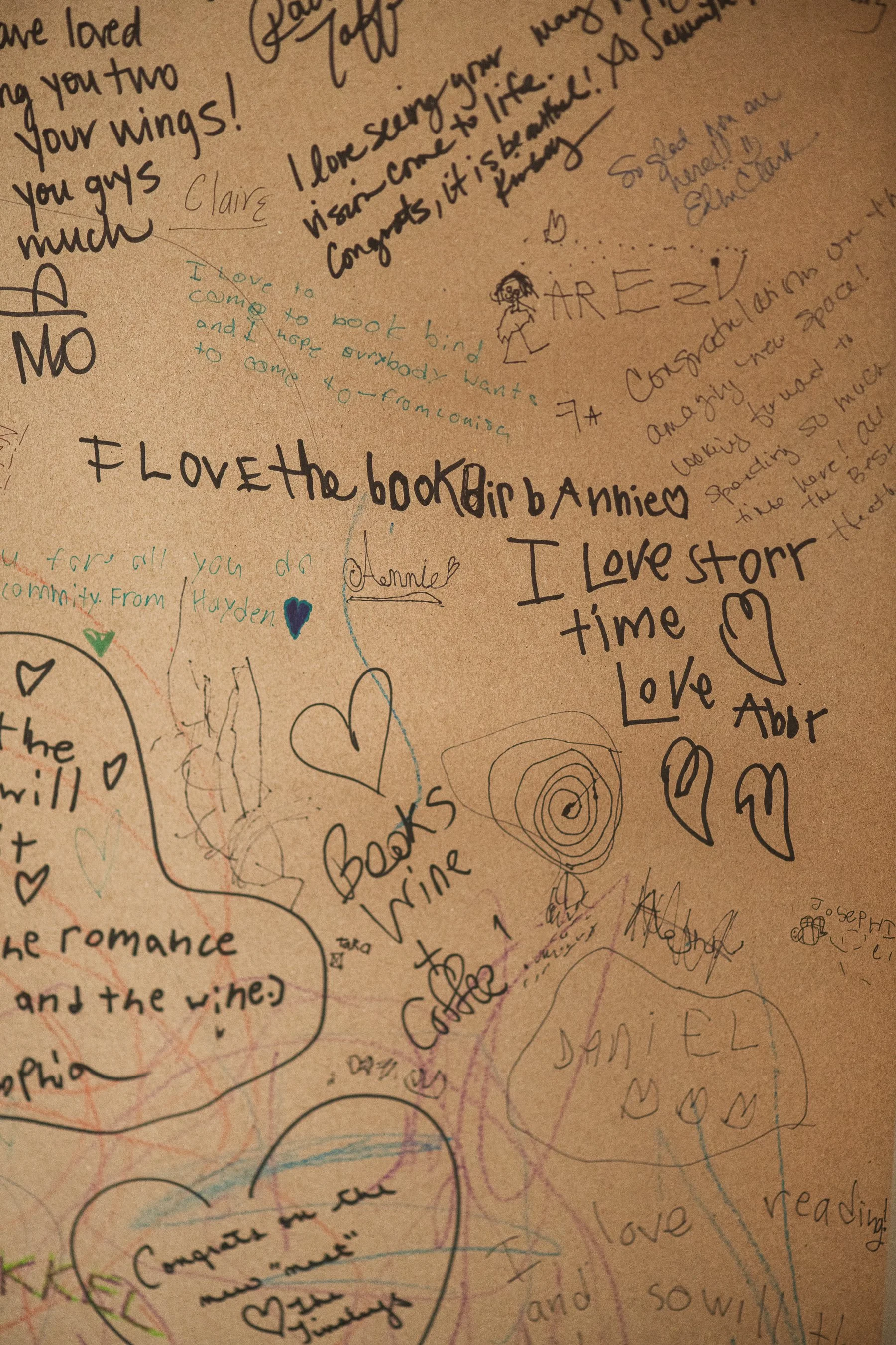 A cardboard surface covered in handwritten notes, messages, and doodles in various colored inks, including hearts, stars, and small drawings.