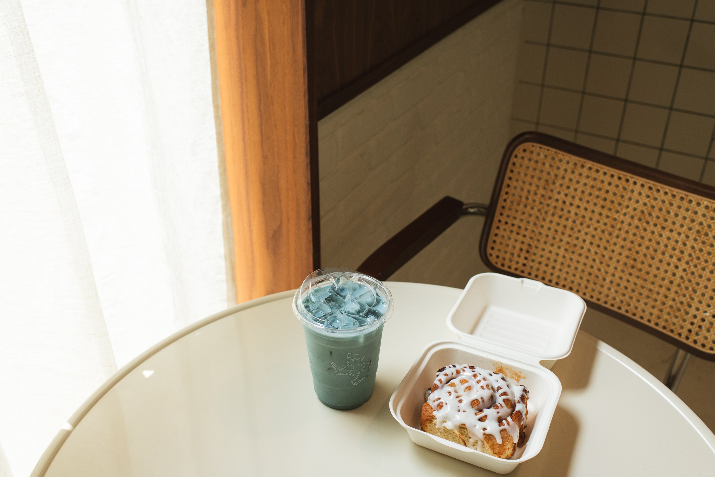 A white high chair with a tray, holding a cup of blue iced drink, a container of cinnamon roll with icing, and a white box. In the background, a brown patterned chair is visible, along with tiled walls and a wooden ledge near a window with beige curtains.