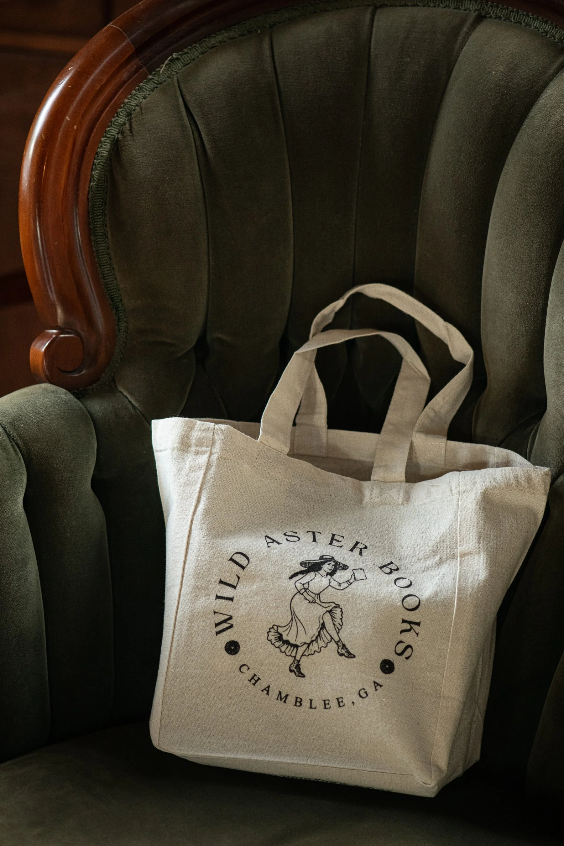 A white tote bag with black text and a drawing of a woman, resting on a dark green upholstered chair with wooden armrest.