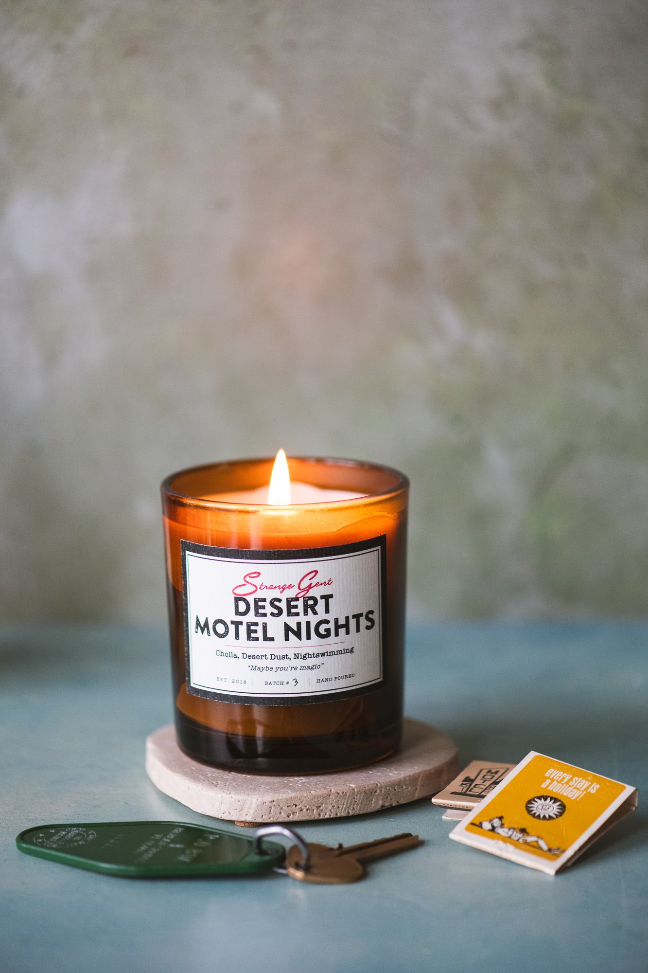 A lit amber glass candle labeled 'Desert Motel Nights' on a round stone coaster, with a key and small book or pamphlet nearby, on a green surface with a textured background.