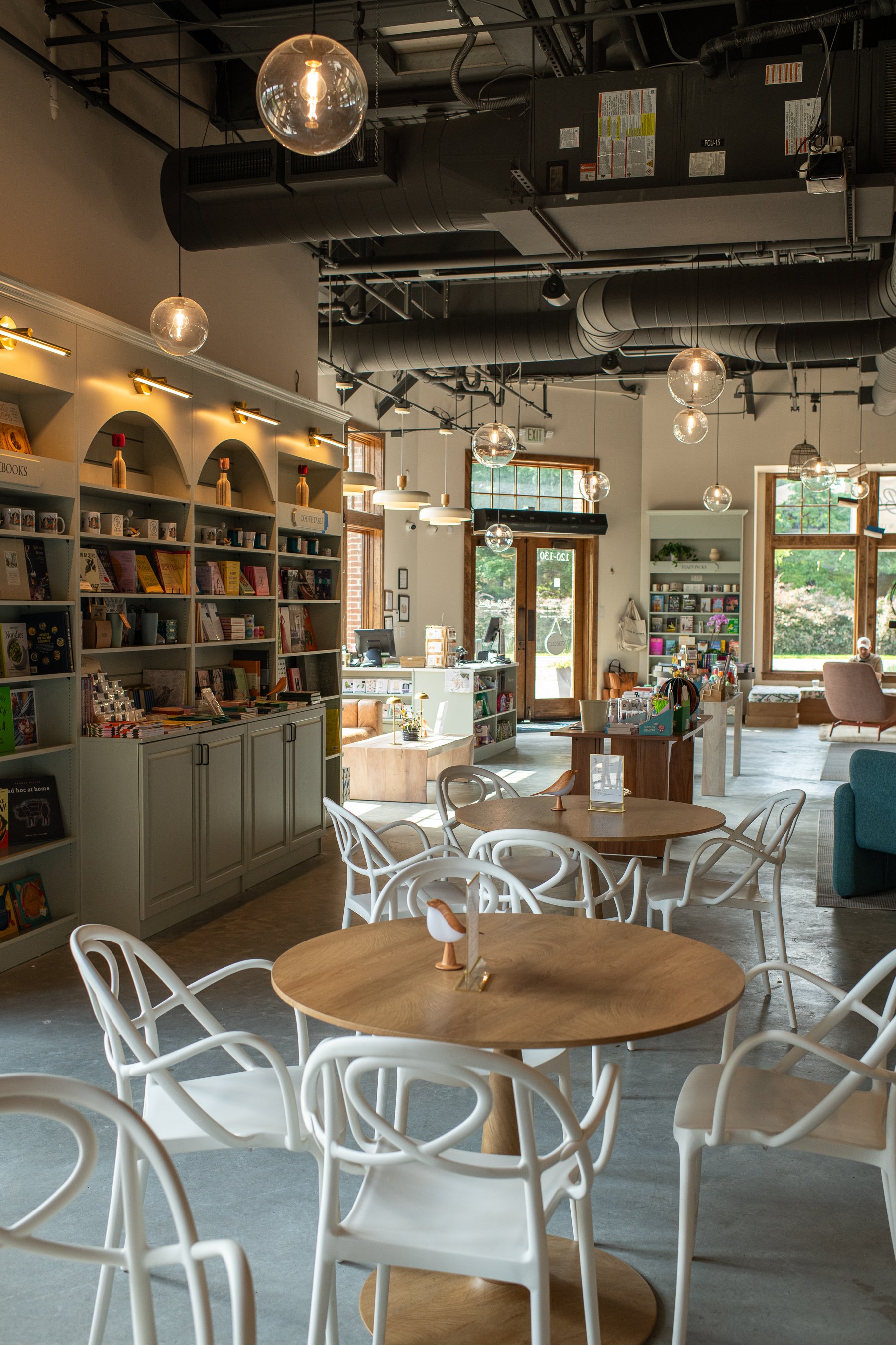 Interior of a modern cafe or bookstore with round wooden tables and white chairs, bookshelves filled with books and decor, large windows with wooden frames, hanging pendant lights, and ceiling ducts.