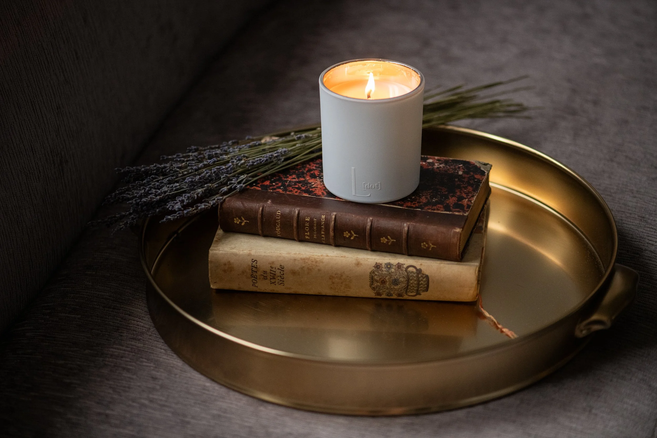 A lit white candle on top of two vintage books inside a gold-colored tray, with dried lavender stems beside the books, on a dark, textured surface.