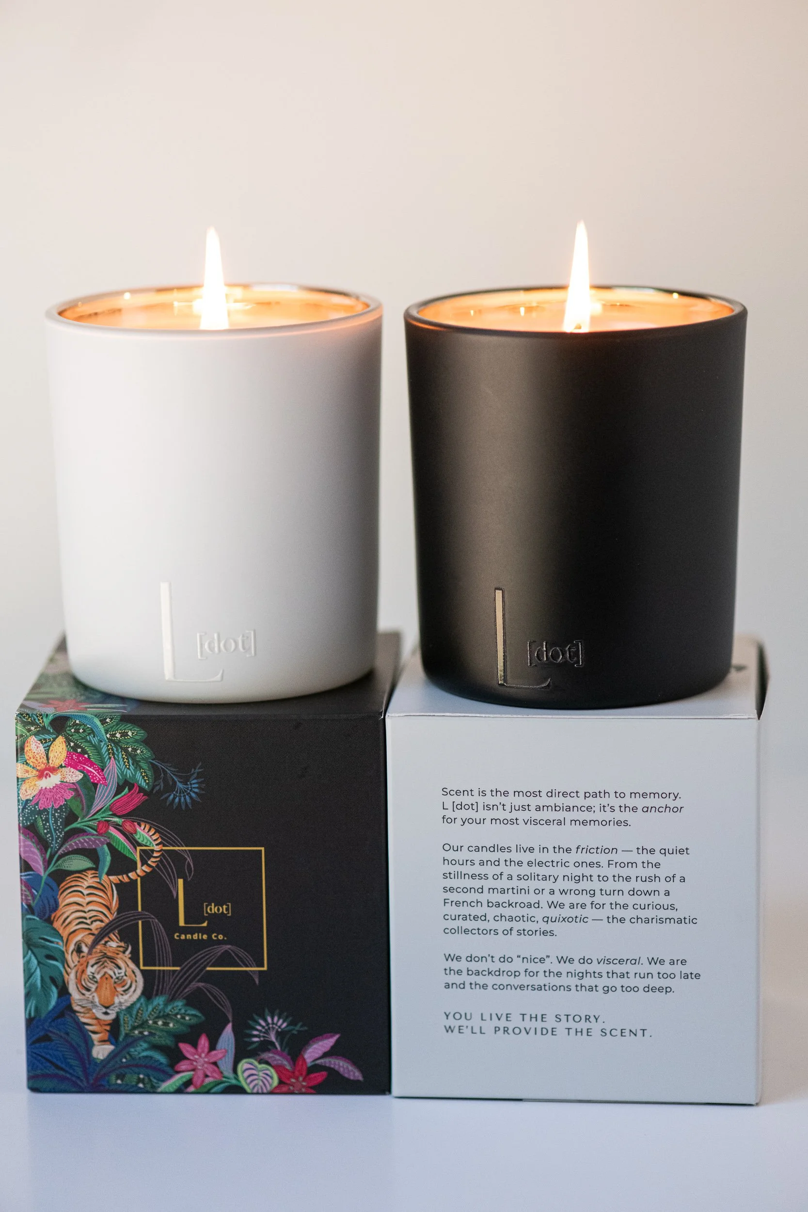Two scented candles, one white and one black, lit and placed on top of decorative boxes. The white box has floral and tiger print design, while the black box has colorful floral artwork. The text on the black box discusses the importance of scent and memory.