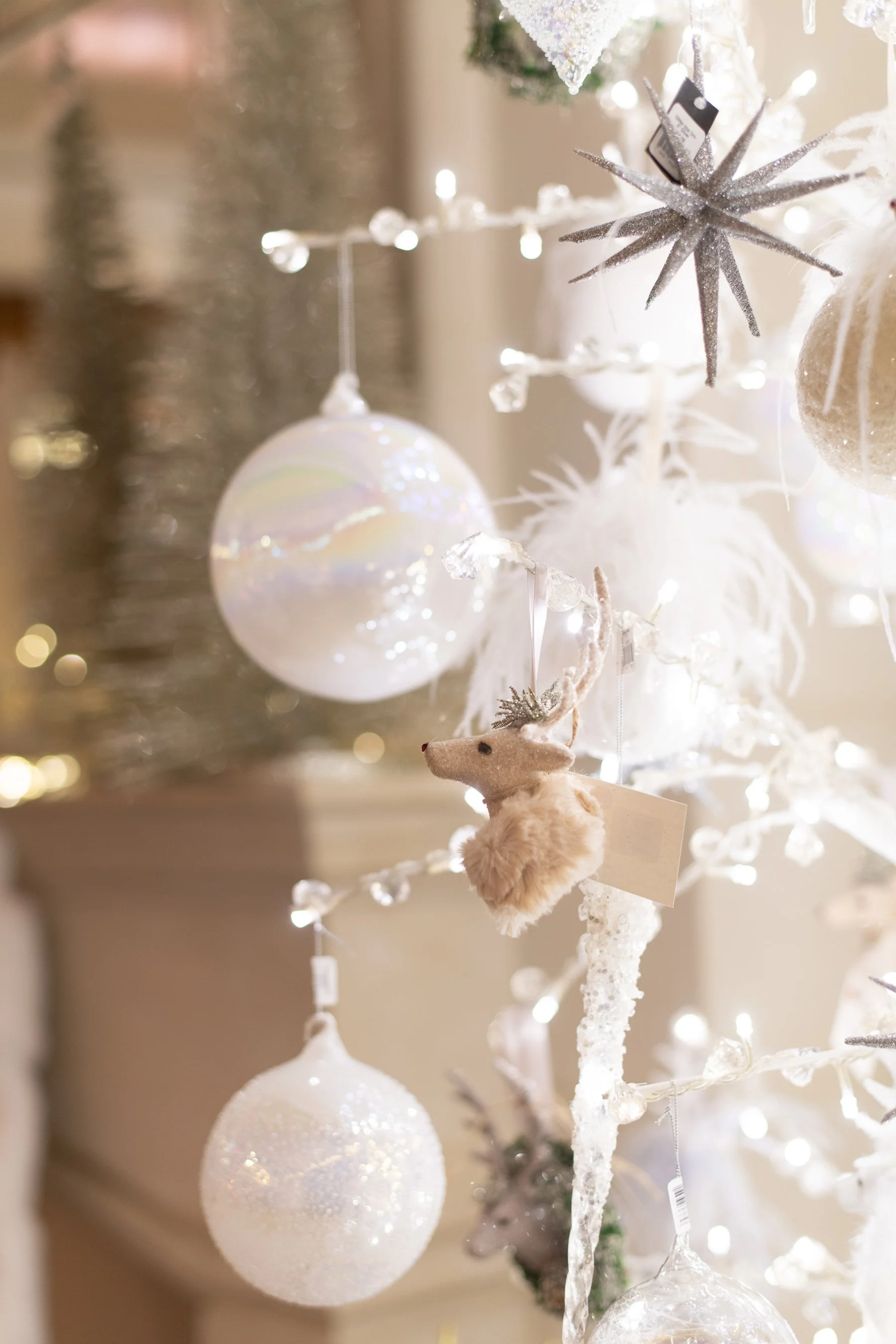 Close-up of a white Christmas tree decorated with white and silver ornaments, including a star-shaped ornament, round baubles, and a small plush reindeer.