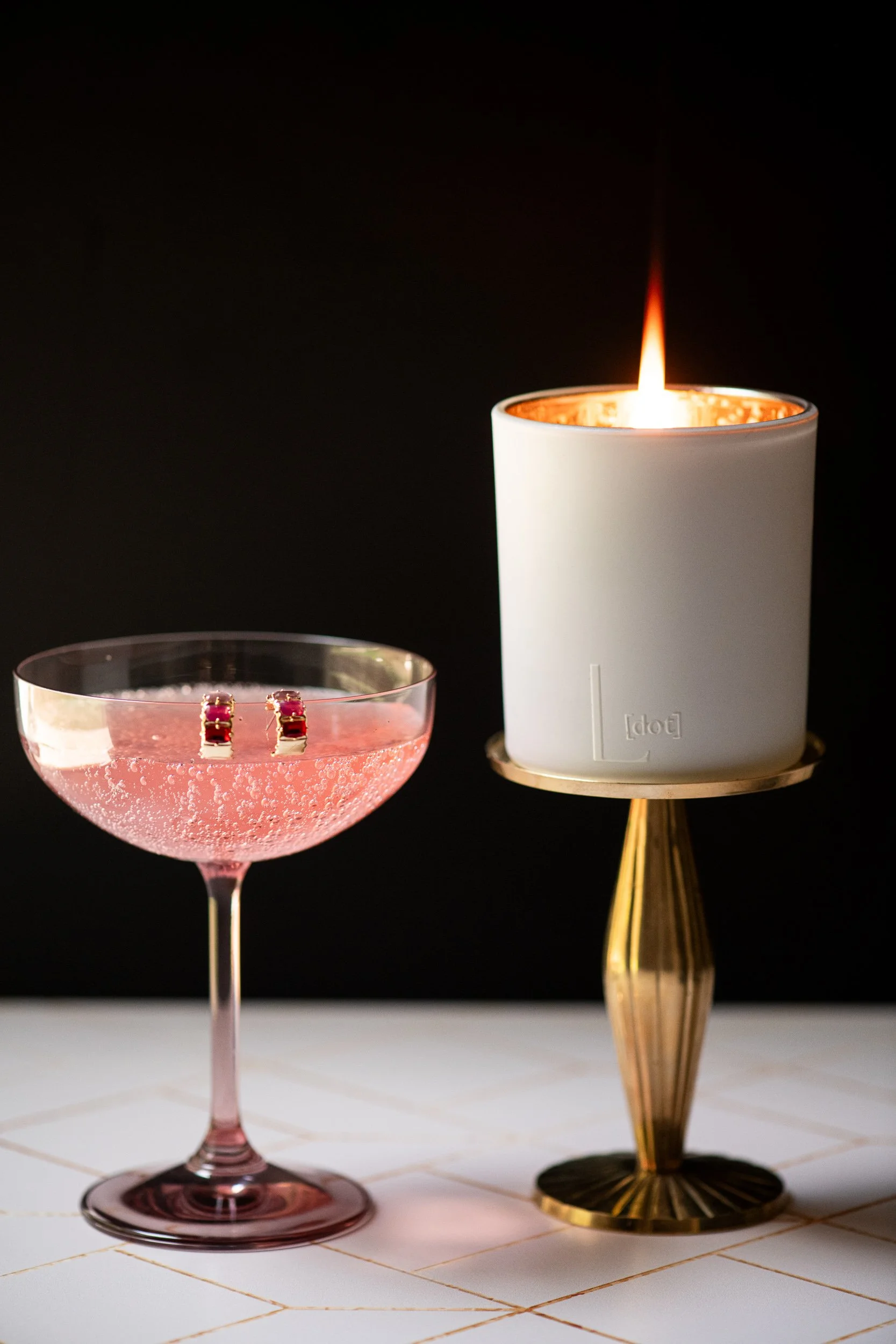 A pink cocktail in a coupe glass with a pair of red gemstone earrings resting inside, and a white candle with an orange flame on a gold stand beside a gold-colored object on a tiled surface against a black background.