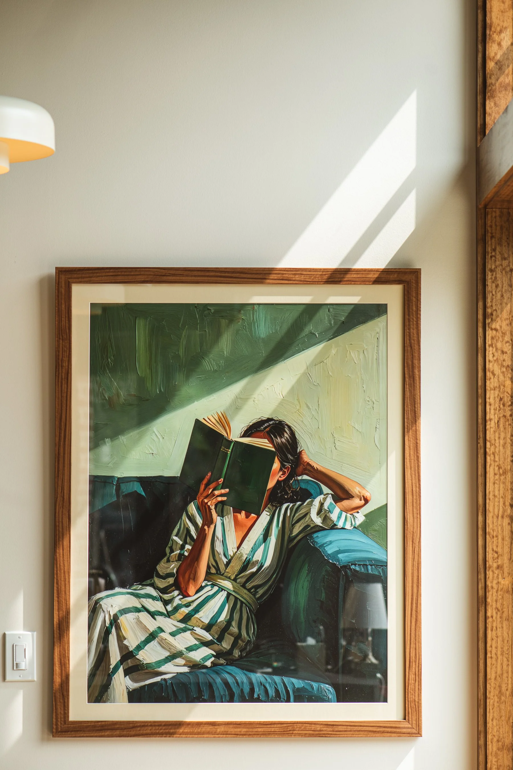 A woman with dark hair wearing a striped dress is sitting in a green armchair and reading a book, with sunlight casting shadows on the wall