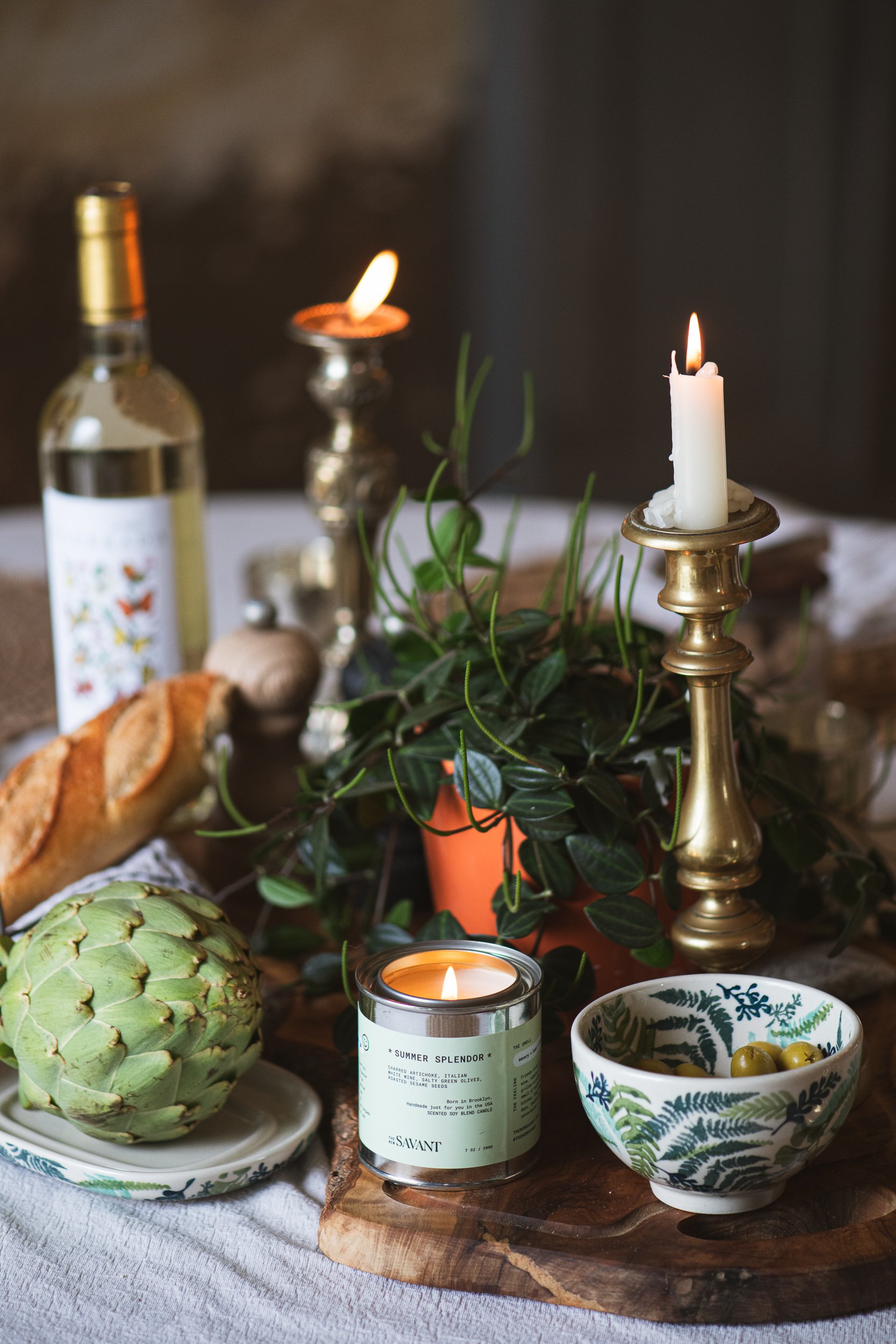 A table setting with candles, a bottle of white wine, artichoke, bowl of green olives, bread, and greenery, arranged on a wooden board and a white tablecloth.