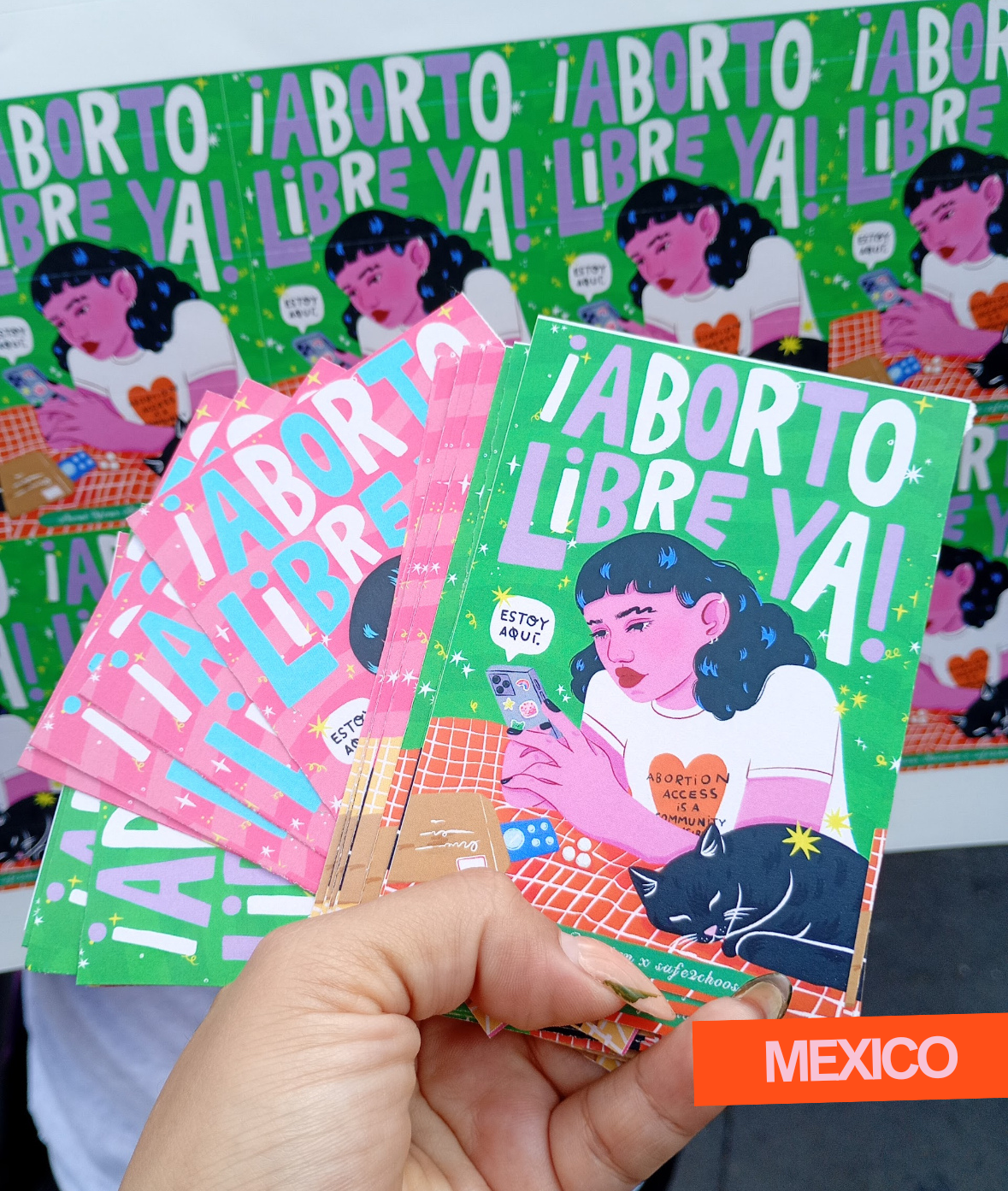 A person holding pamphlets with a woman looking at her phone on the cover, with a black cat sleeping beside her. The pamphlet has the text '¡Aborto Libre Ya!' and 'Estoy Aquí!' in Spanish, indicating support for abortion rights. The background has similar posters and there's an orange banner in the corner with the word 'Mexico'.