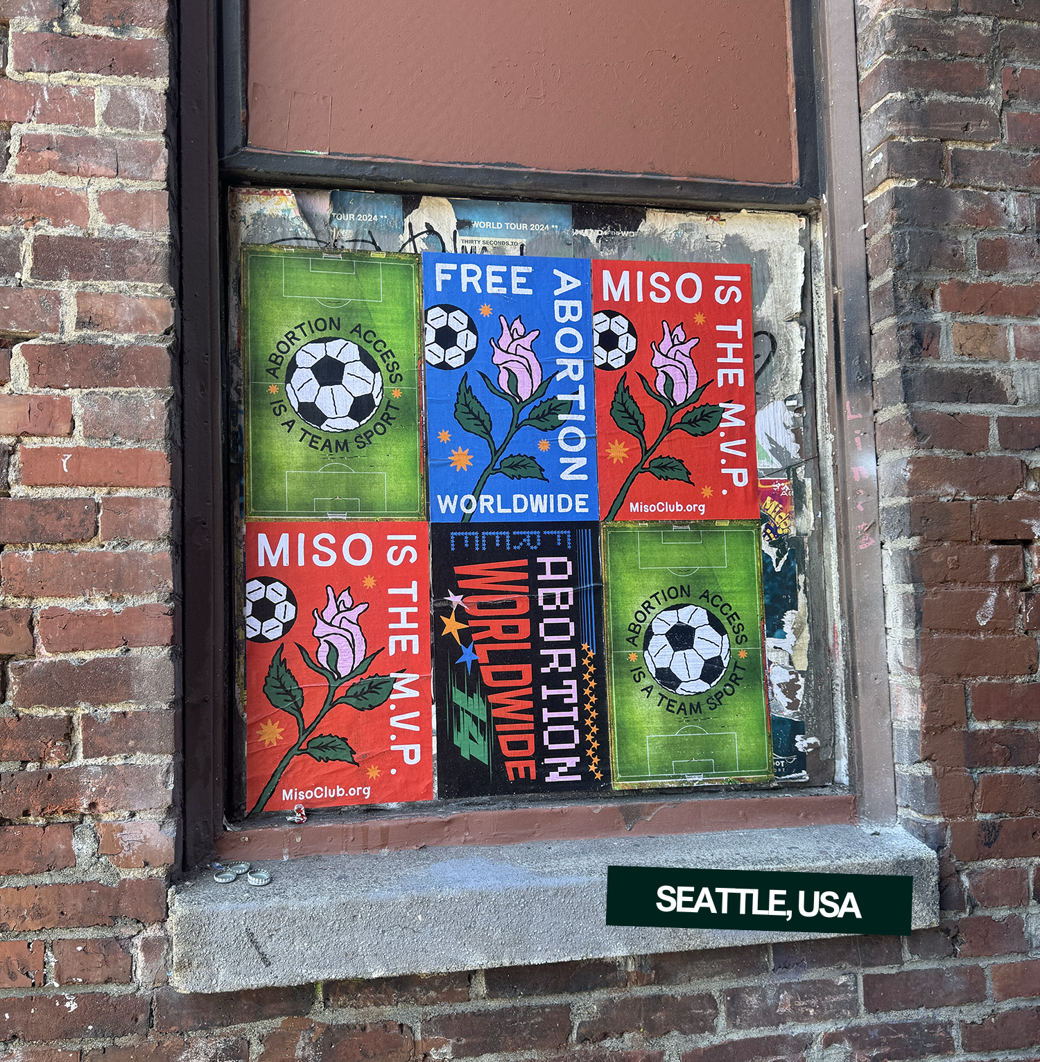 A display window with stickers on it, including two green soccer field designs with the words 'ABORTION ACCESS' and 'A TEAM SPORT,' two blue and red stickers promoting free abortions worldwide, featuring a pink flower and a soccer ball, and other stickers with the text 'IS THE M.V.P.' and 'ABORTION WORLDBOWL.' The window is surrounded by a brick wall, and a sign reading 'SEATTLE, USA' is below the window.